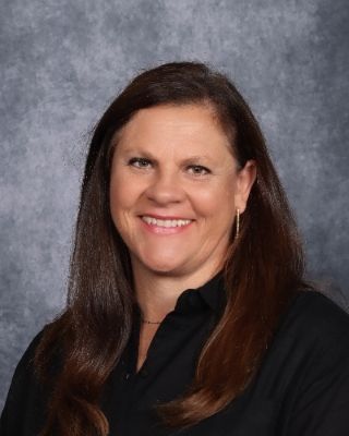Woman with brown hair, smiling, wearing a black shirt, in front of a gray-blue backdrop.