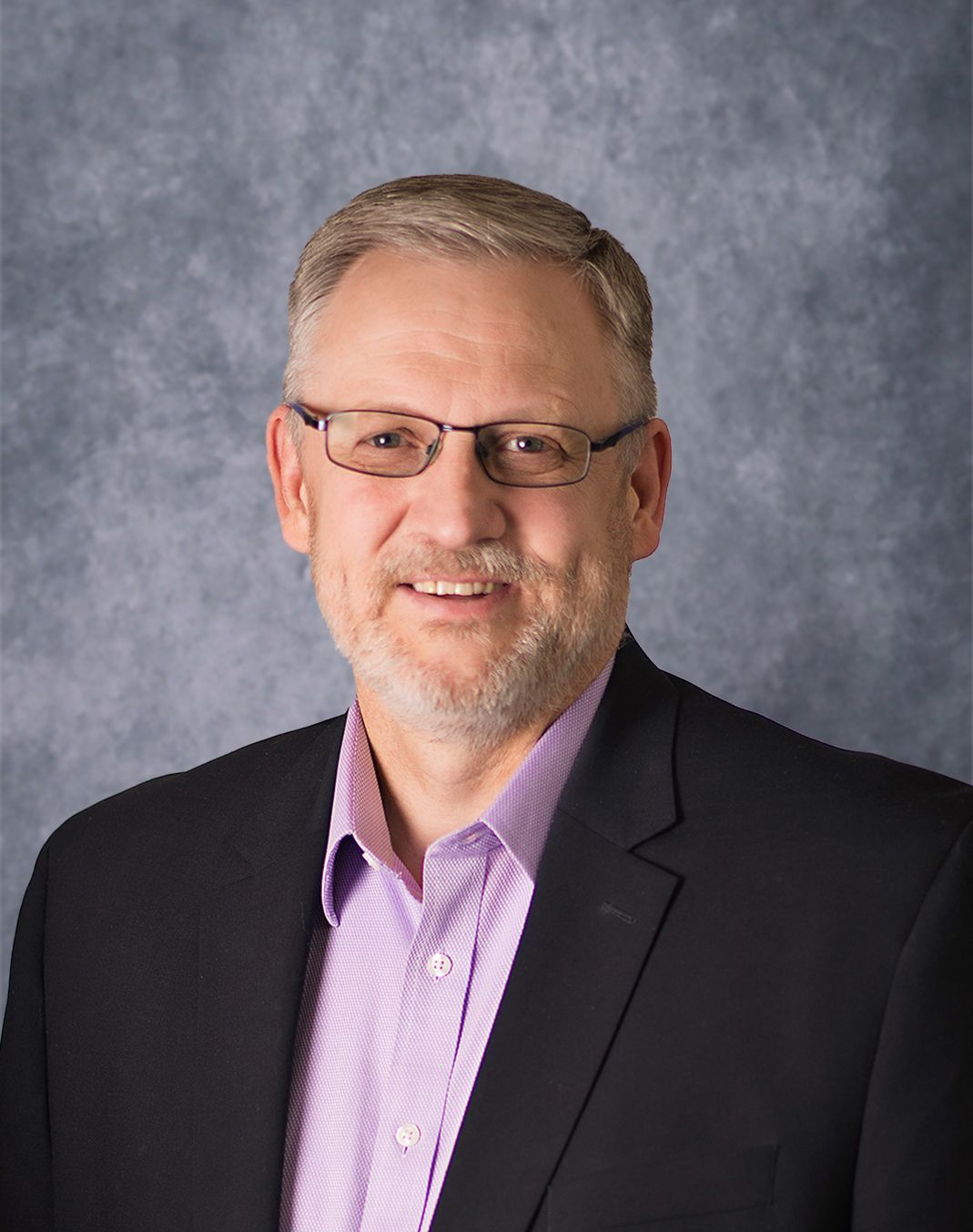 Man with glasses and beard in a suit jacket, purple shirt, smiling, against a blurred gray background.