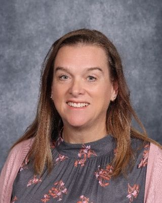 Woman with long brown hair smiles, wearing a gray floral top and pink cardigan against a gray background.