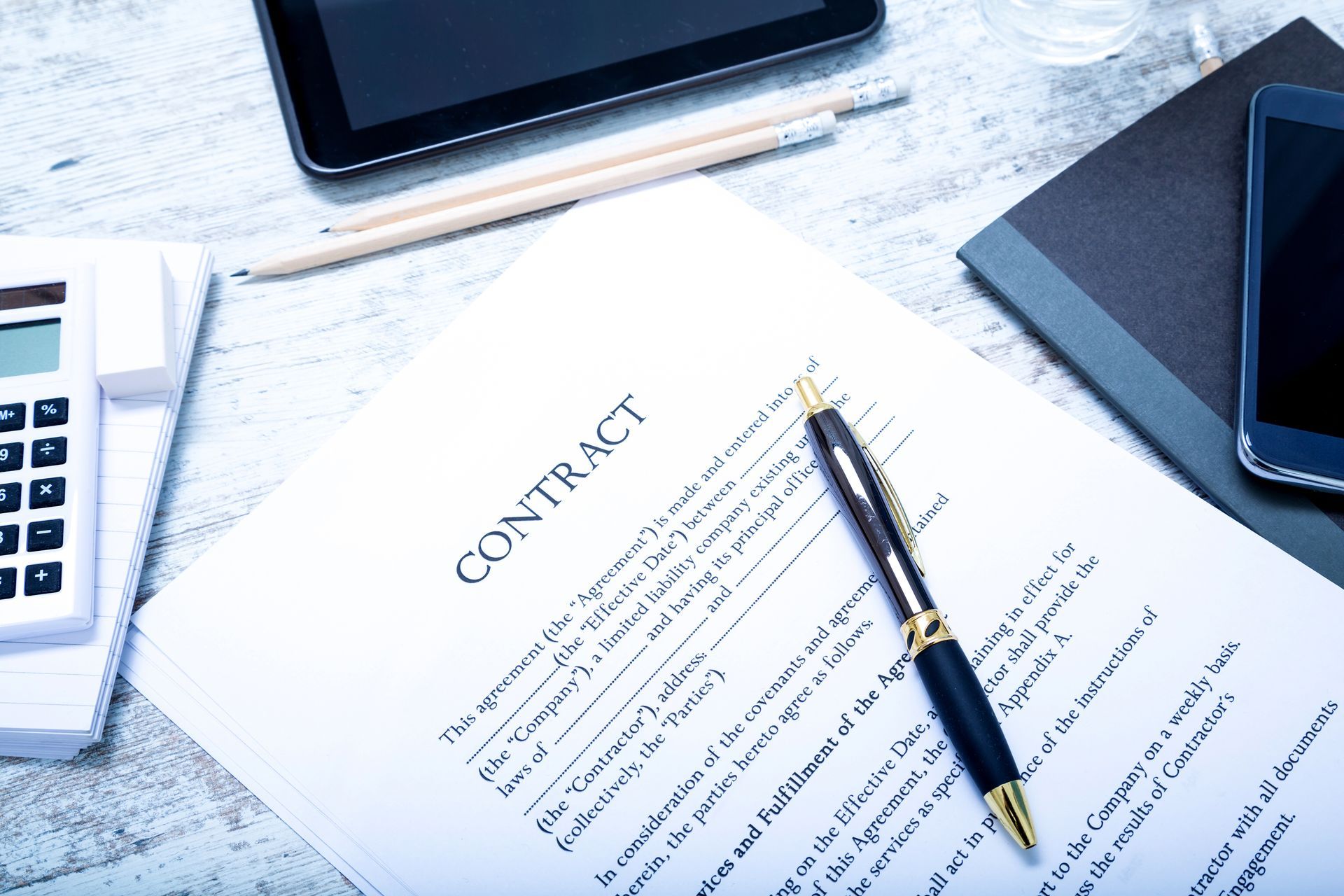 Contract document with pen, tablet, phone, calculator, and notebook on a white wooden surface.