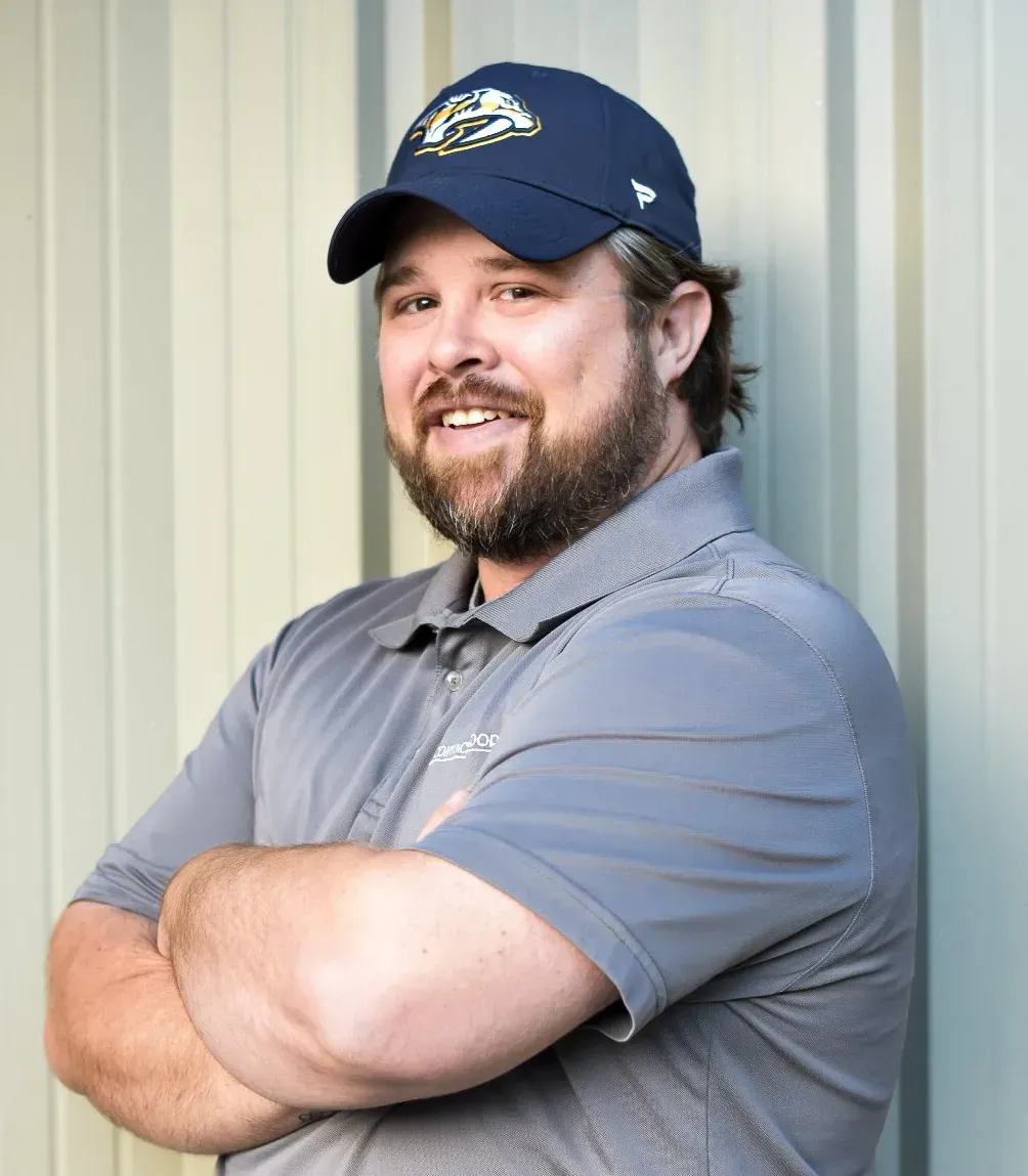 A man wearing a blue hat and a grey shirt is standing with his arms crossed.