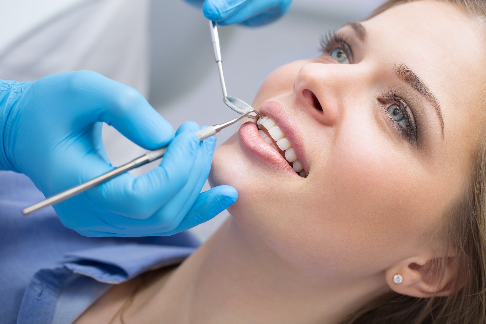 Dentist examining a woman's teeth with tools in a dental office. Blue gloves and patient smiling.