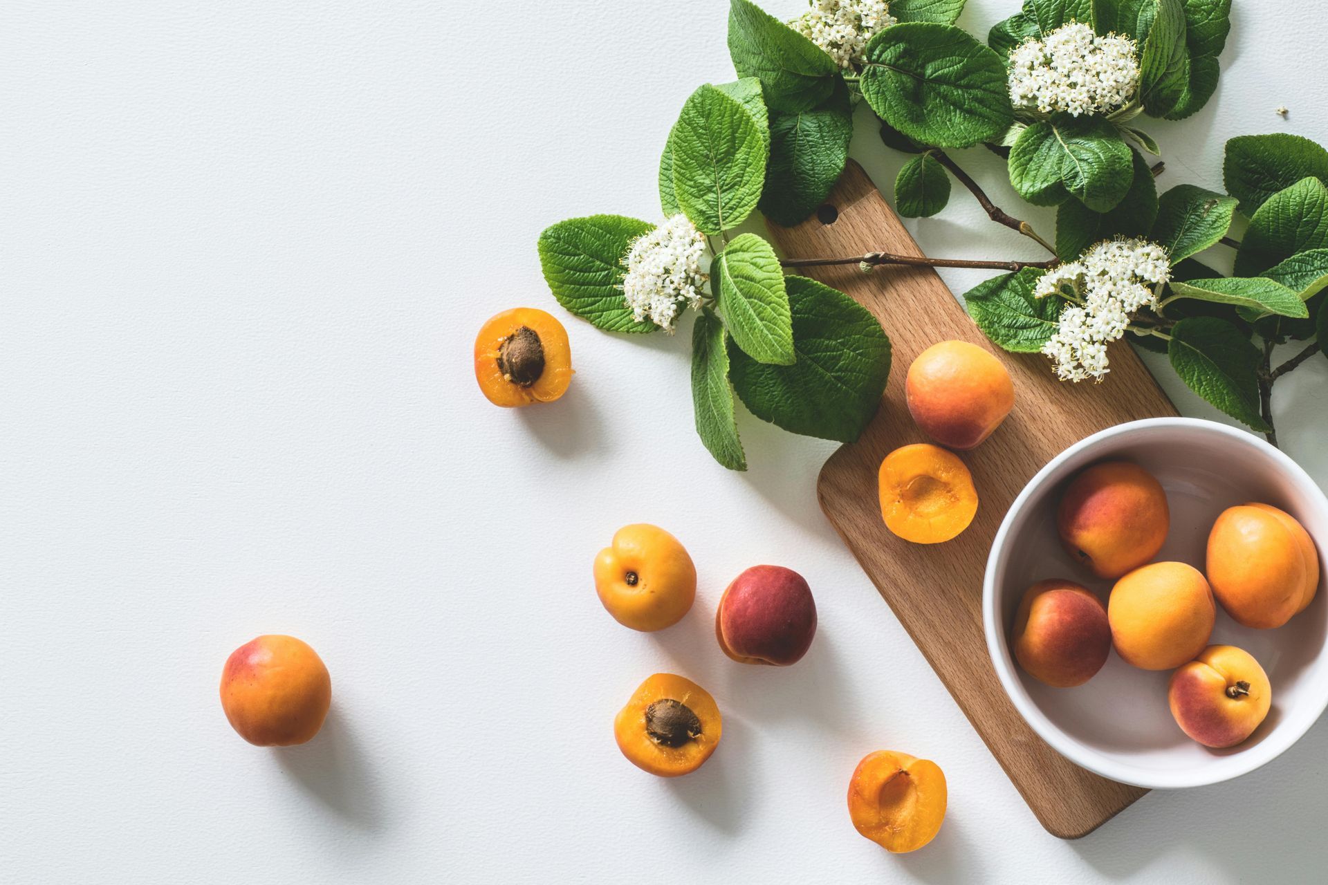 Apricots on a wooden board and in a white bowl with green leaves and white flowers, on a white surface.