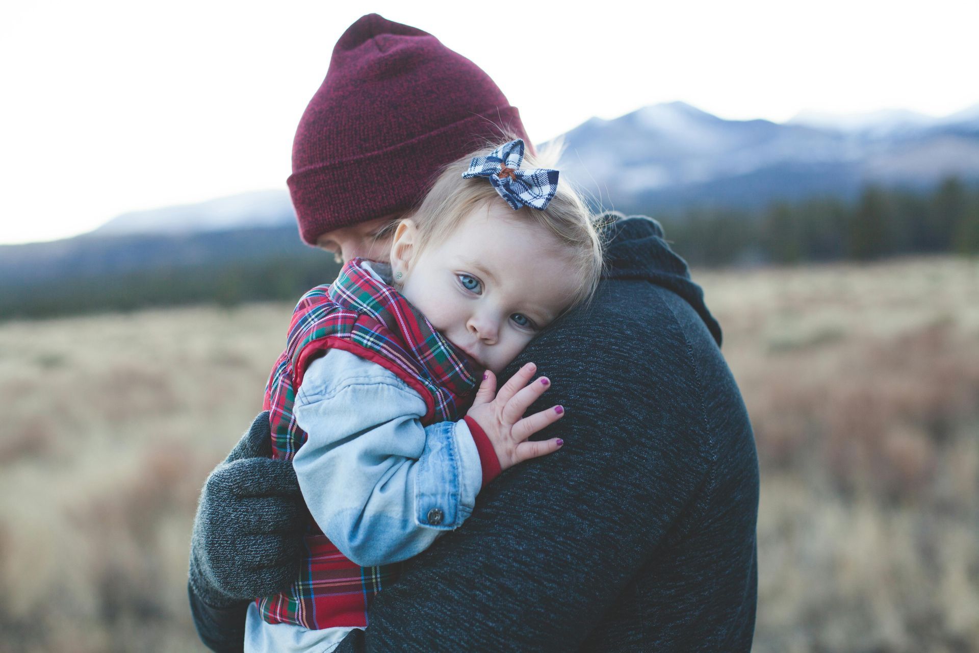 Person holding a baby outdoors. Both are bundled up; the baby looks at the camera. Mountains in background.