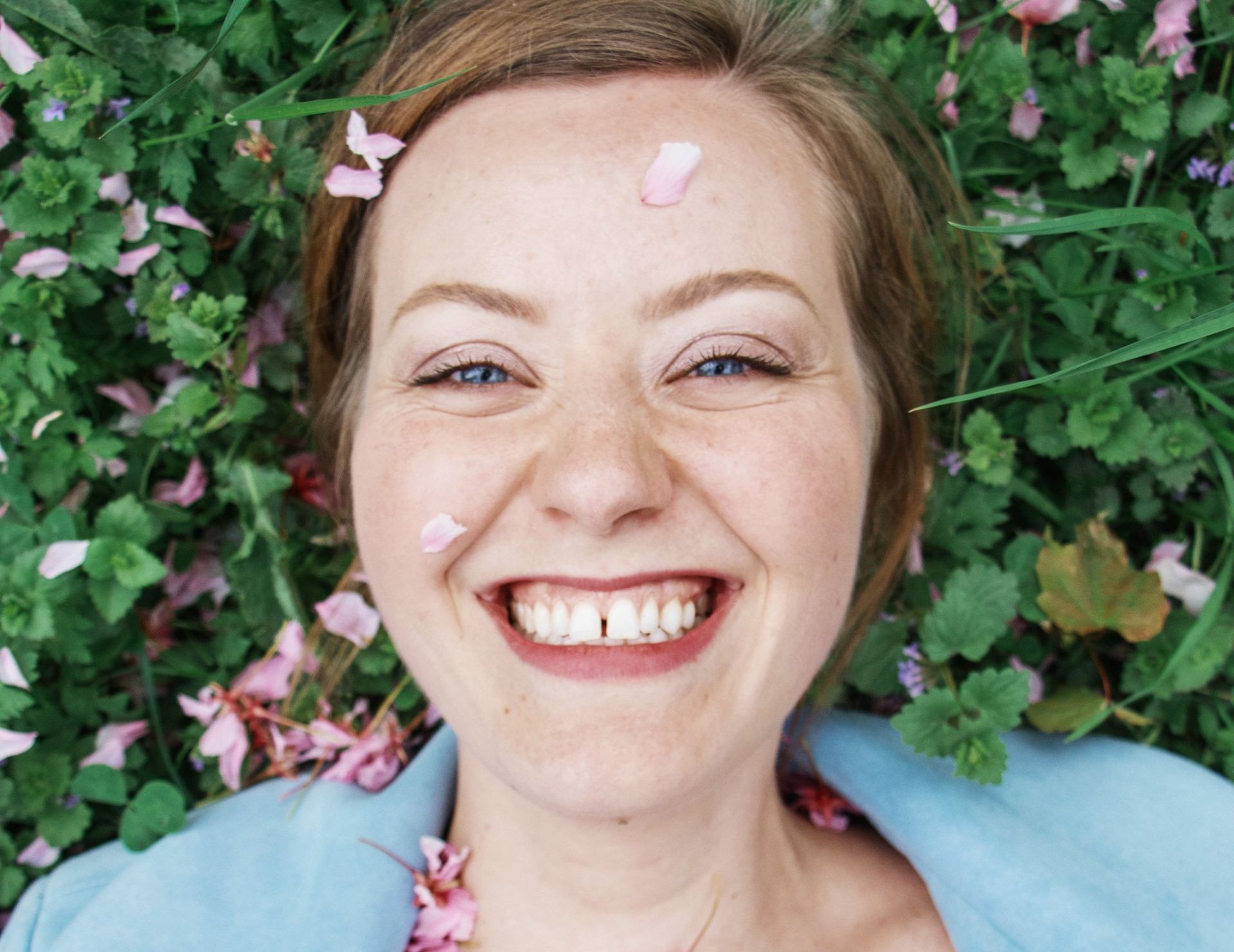 A woman is laying in the grass with flowers on her face and smiling.
