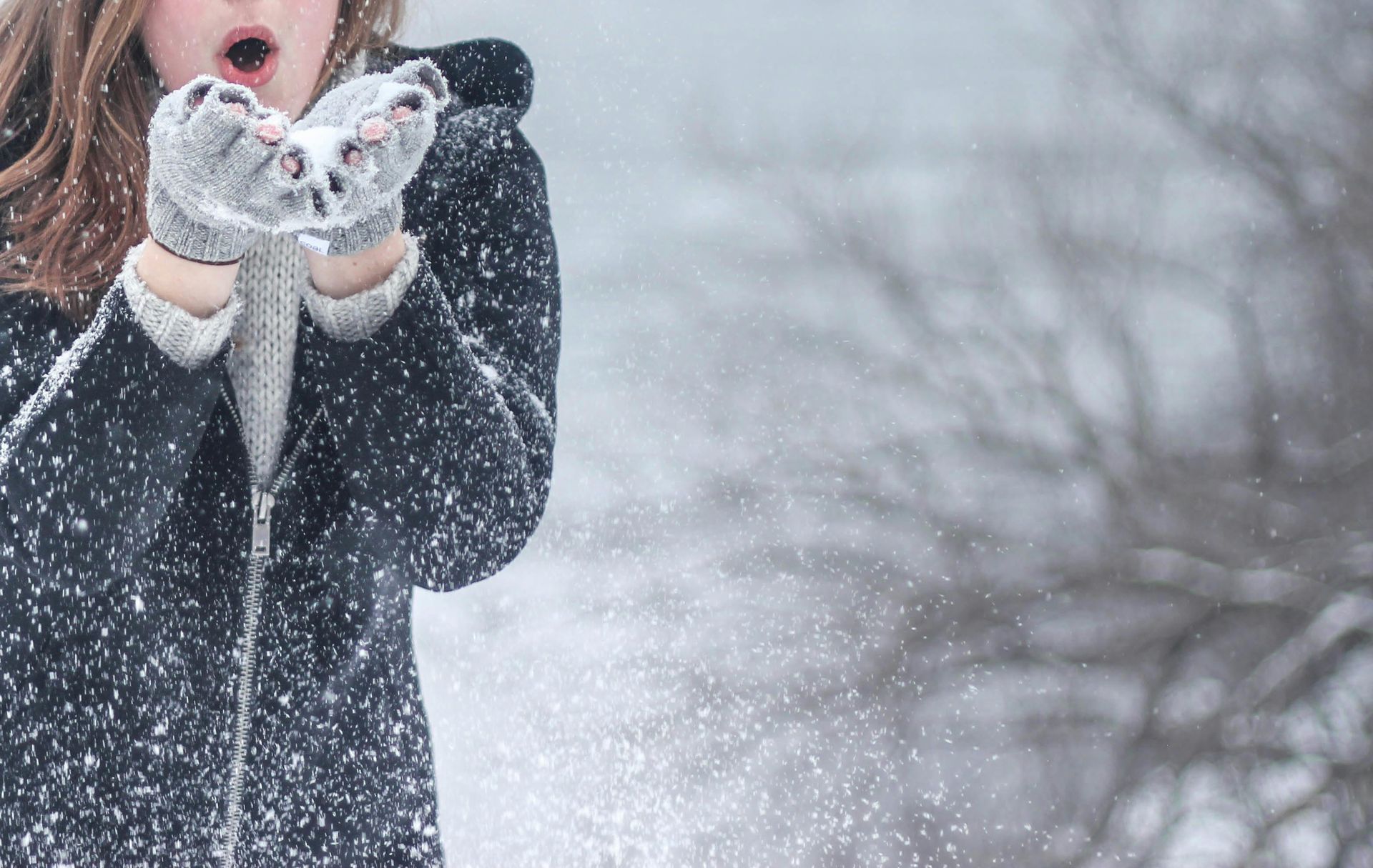 Woman in dark coat blowing snowflakes from gloved hands in a snowy setting.