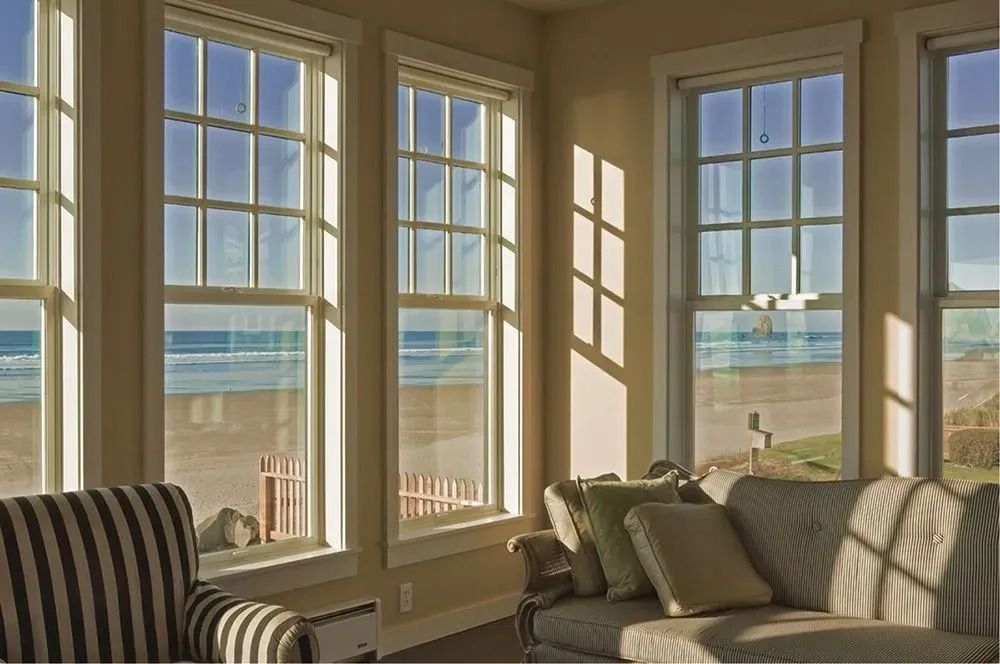 Interior room with tall windows overlooking a beach and ocean. Sunlight streams through the windows.