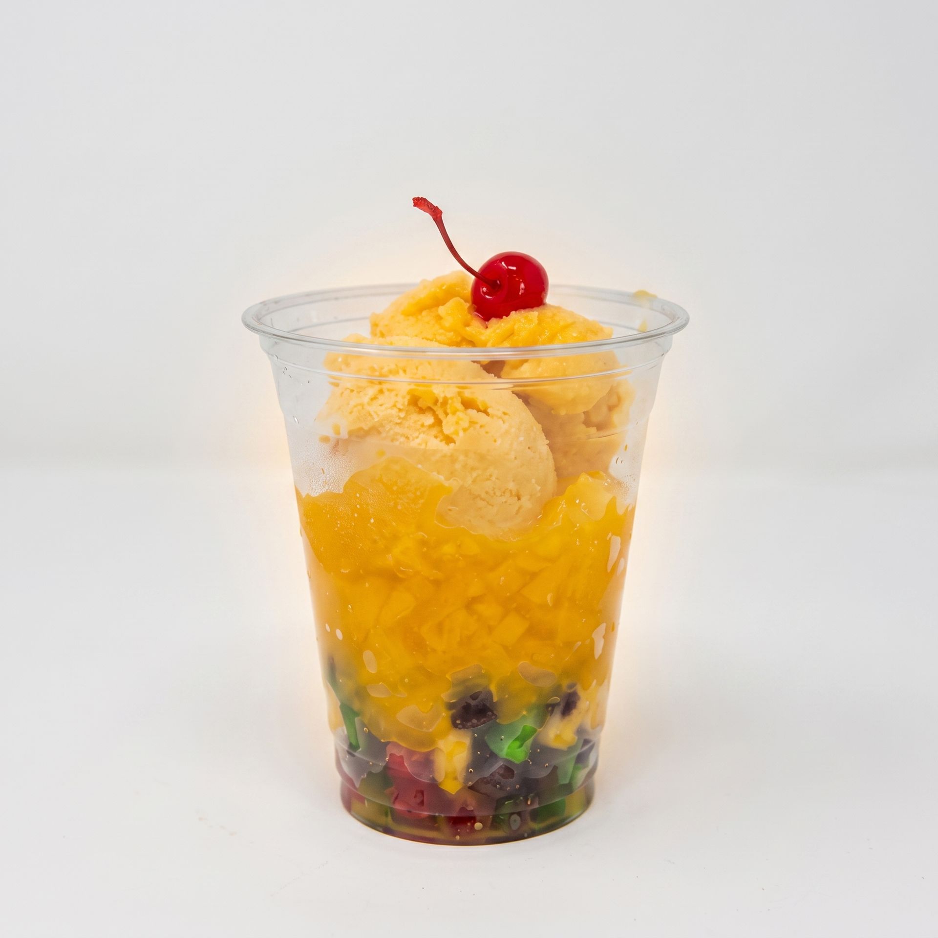 Clear cup of yellow shaved ice topped with a cherry, with fruit and jelly at the bottom.