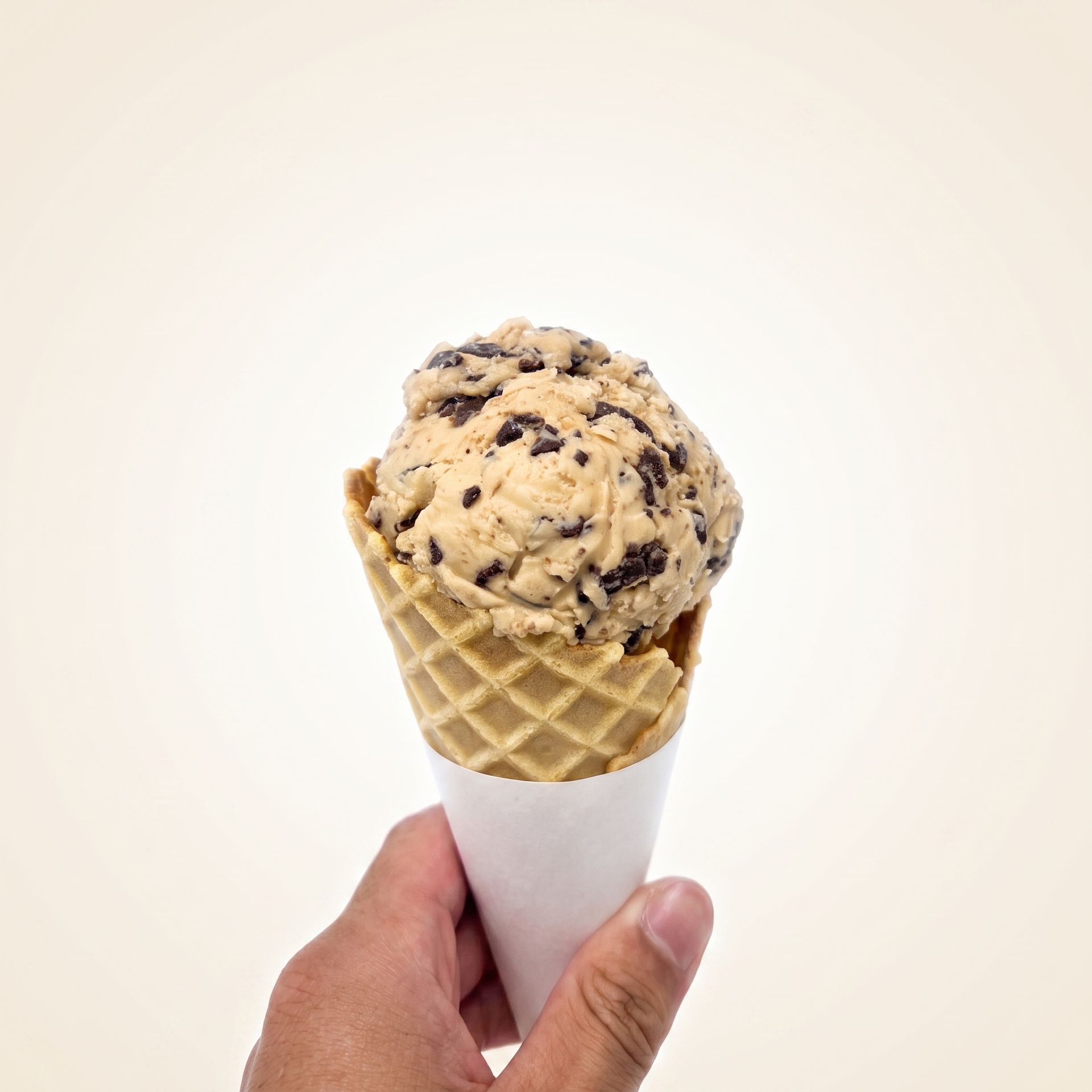 Hand holding a waffle cone with cookie dough ice cream against a plain white background