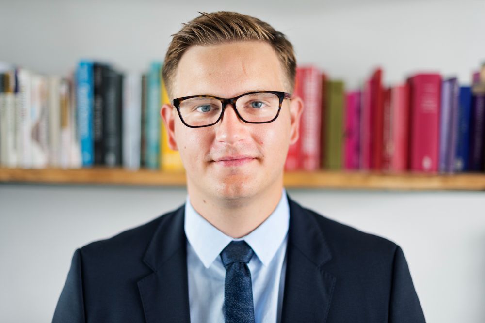 A man in a suit and tie is standing in front of a bookshelf.