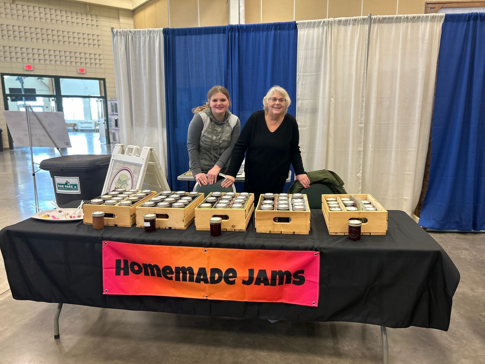 Two women are standing behind a table selling homemade jams.