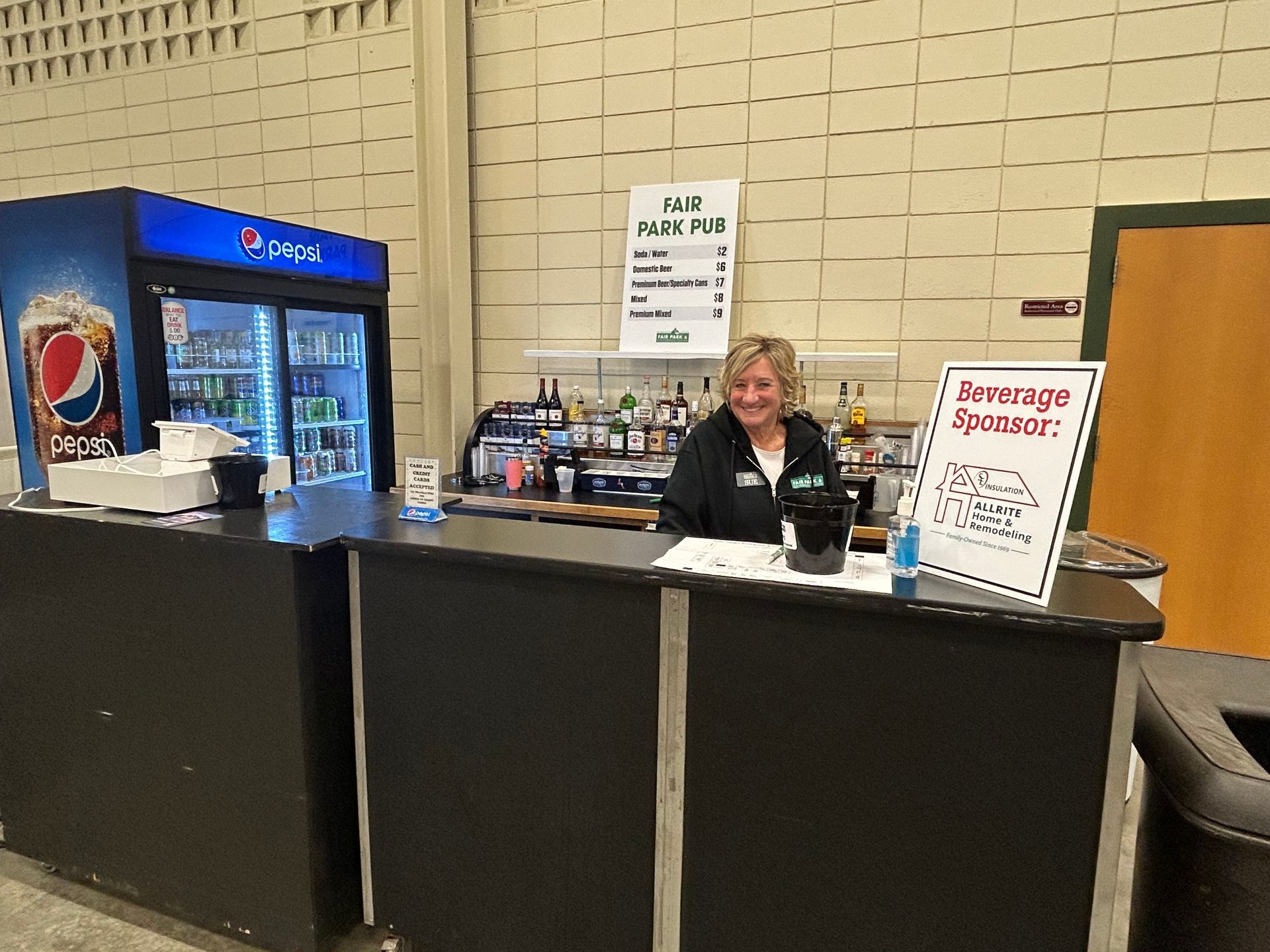 A woman is standing behind a counter in front of a pepsi refrigerator.