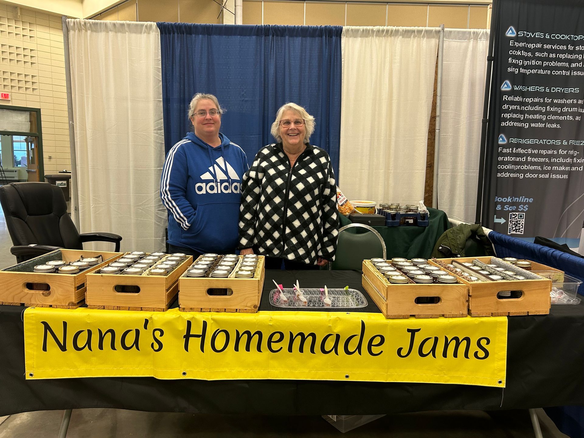 Two women are standing behind a table selling nana 's homemade jams.