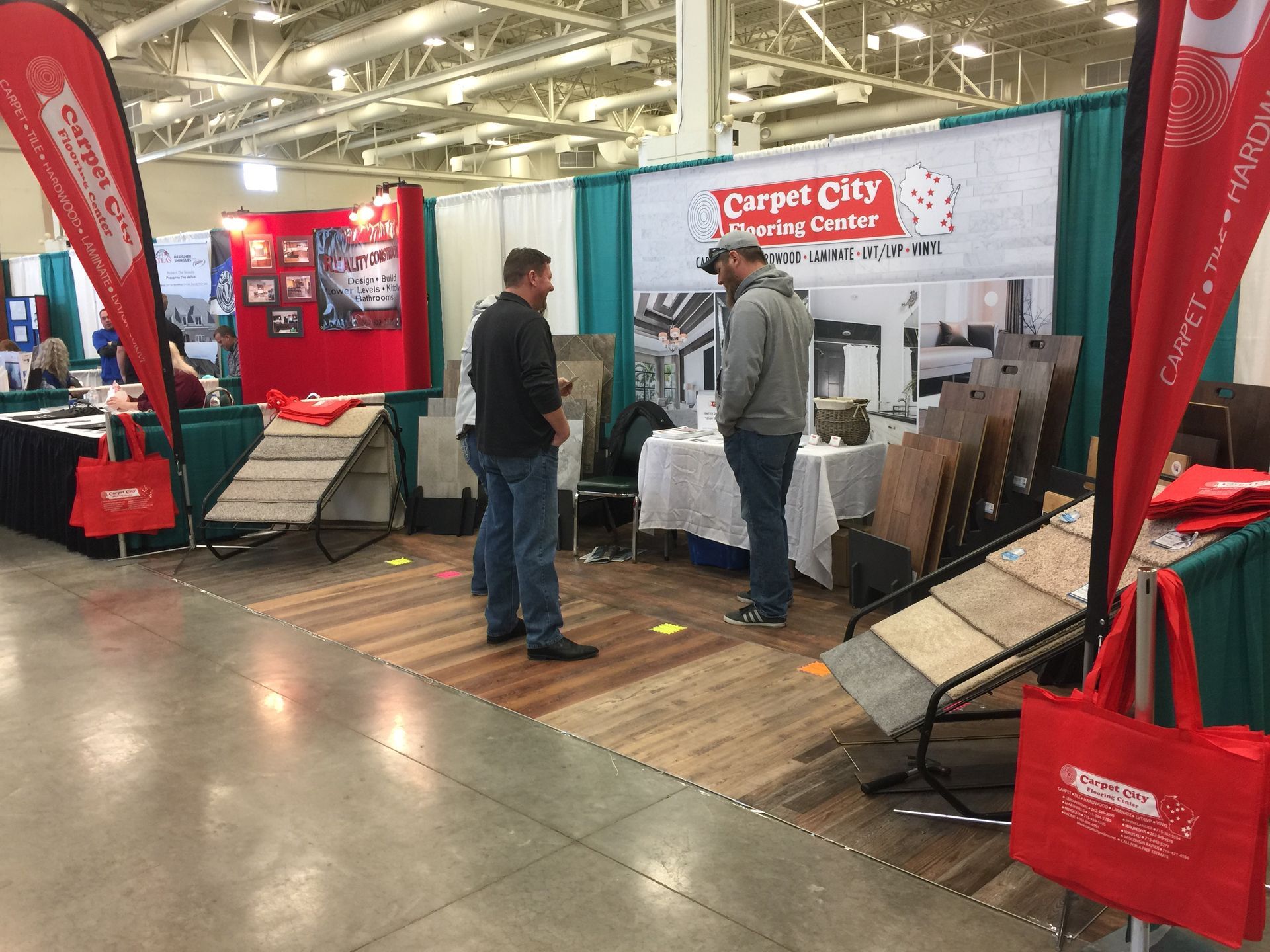 Two men are standing in front of a carpet city booth at a trade show.