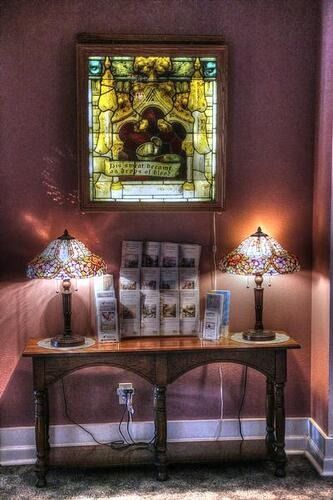 Wooden table with lamps on either side, below stained glass window, brochures displayed.