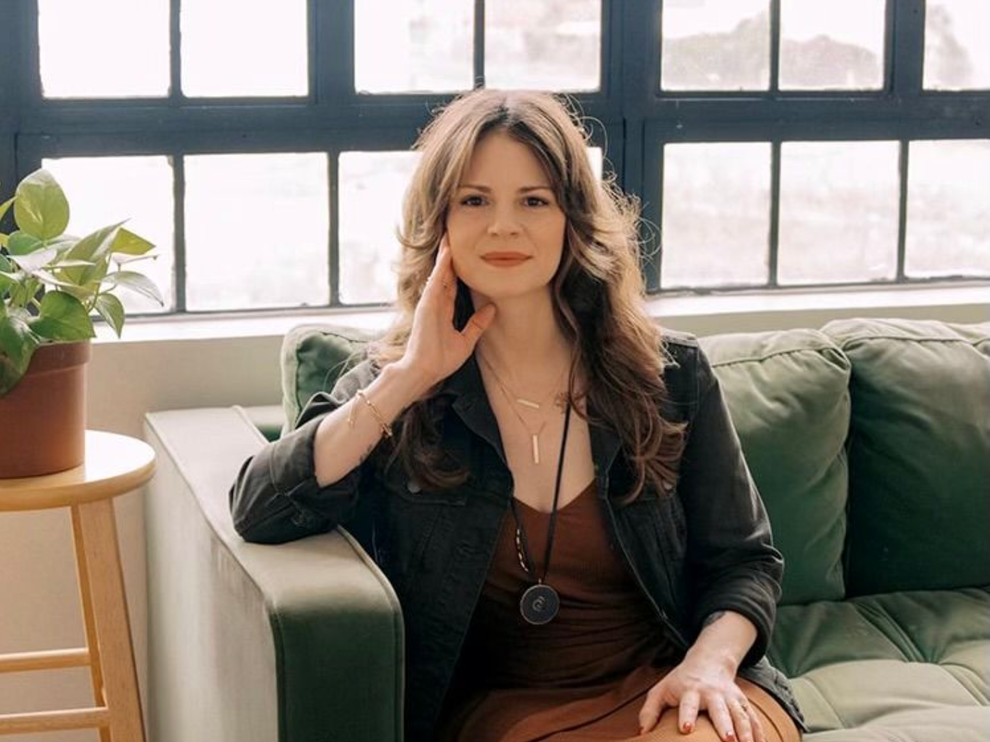 Woman sitting on a green couch by a bright window, hand on face, with a potted plant nearby