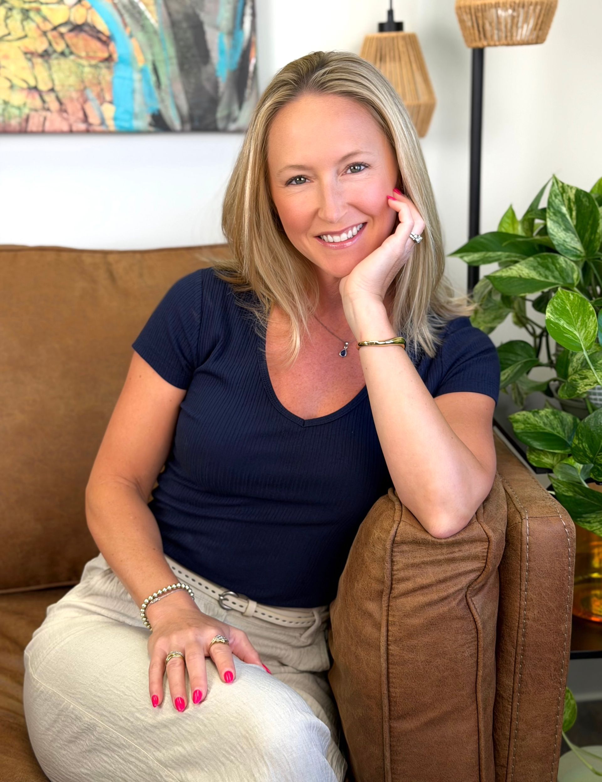 Smiling woman seated on a couch with a hand on her chin, beside a potted plant and framed wall art.