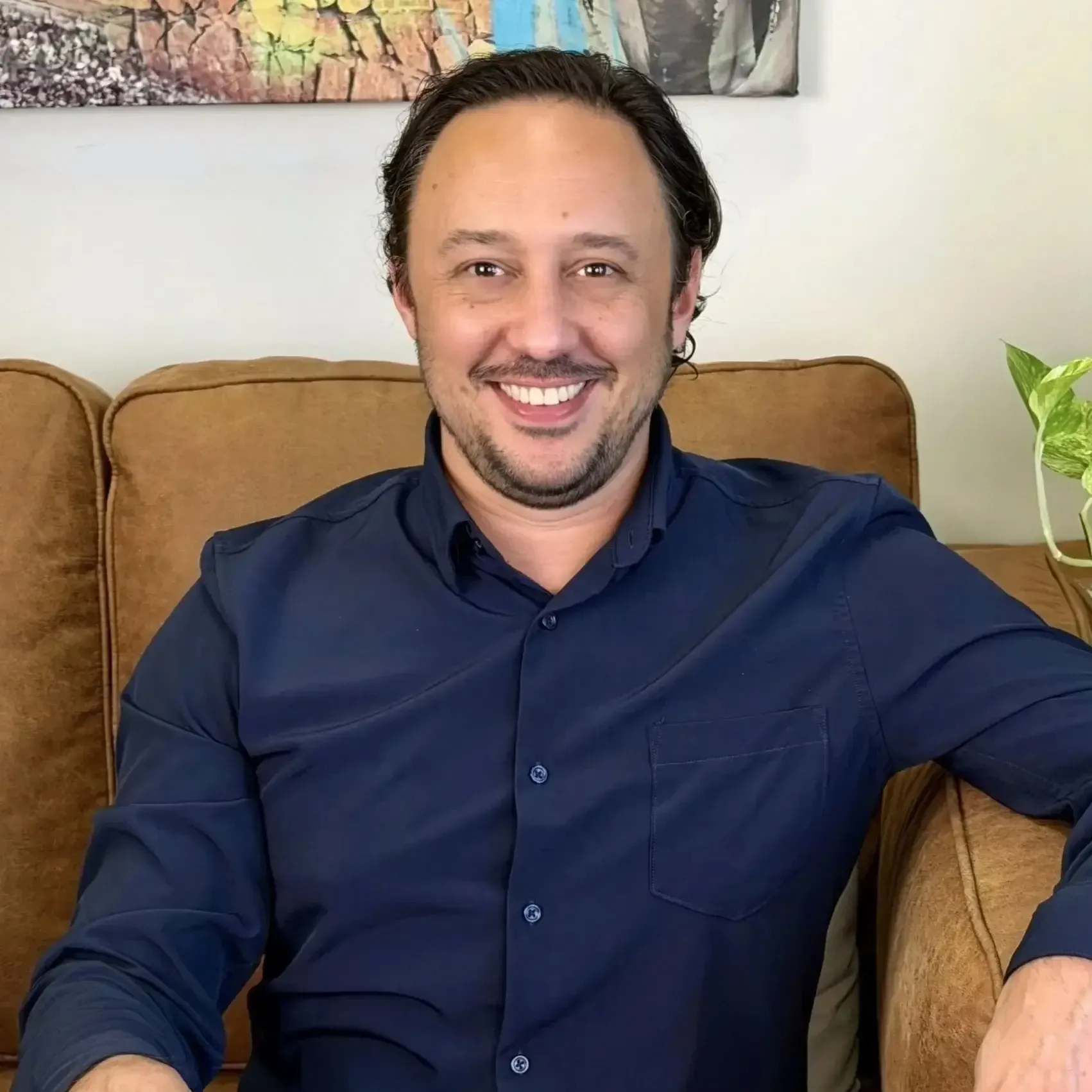 Smiling man in a navy shirt sitting on a tan couch in a bright living room.