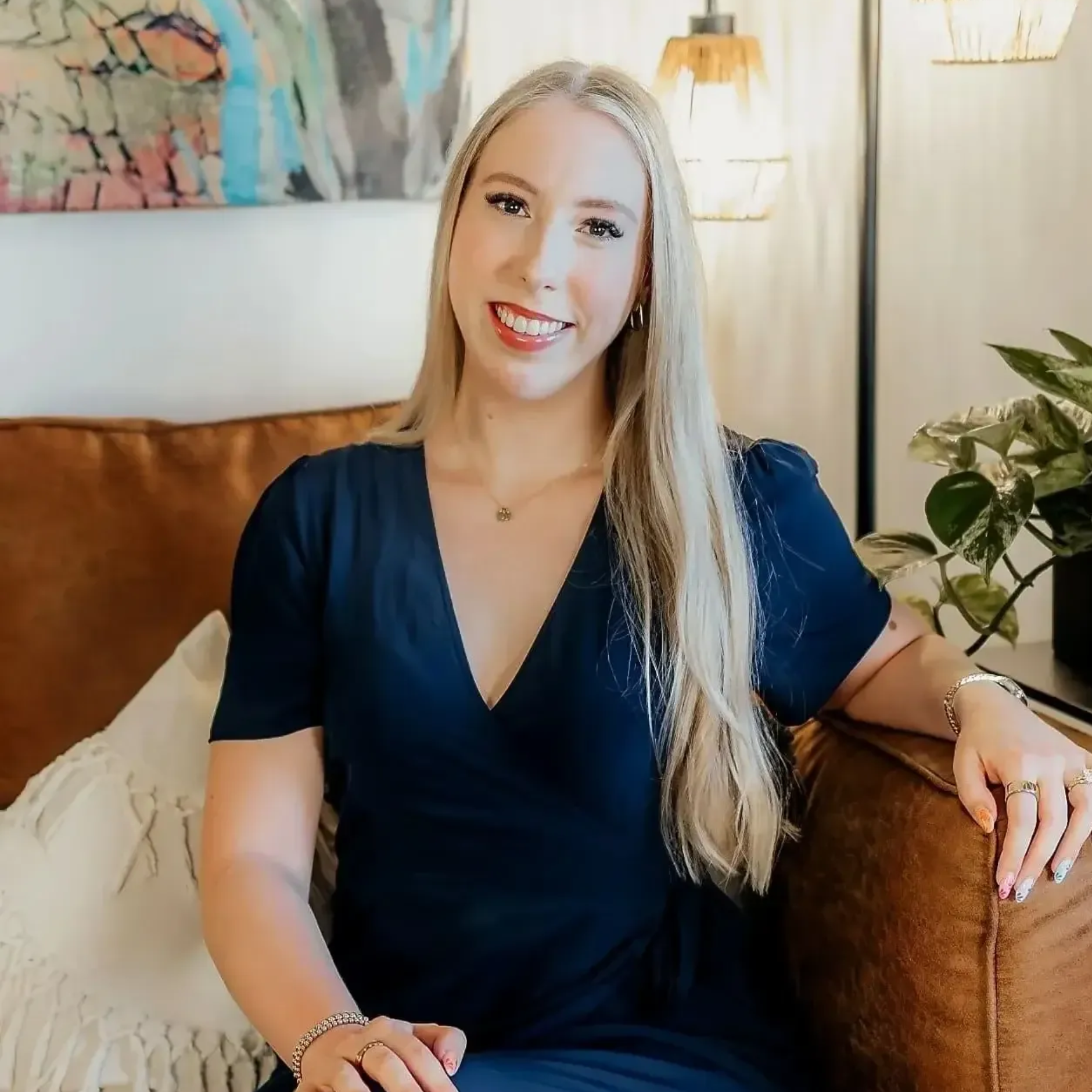 Smiling blonde woman in a navy dress seated on a brown couch in a bright living room