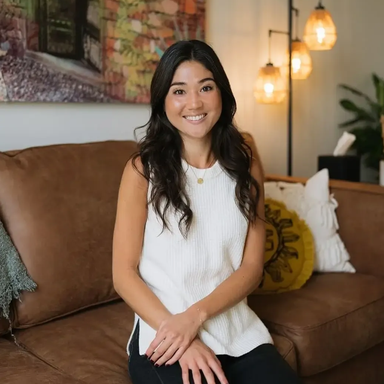 Woman sitting on a brown couch, smiling in a warmly lit living room with hanging lights and patterned decor.