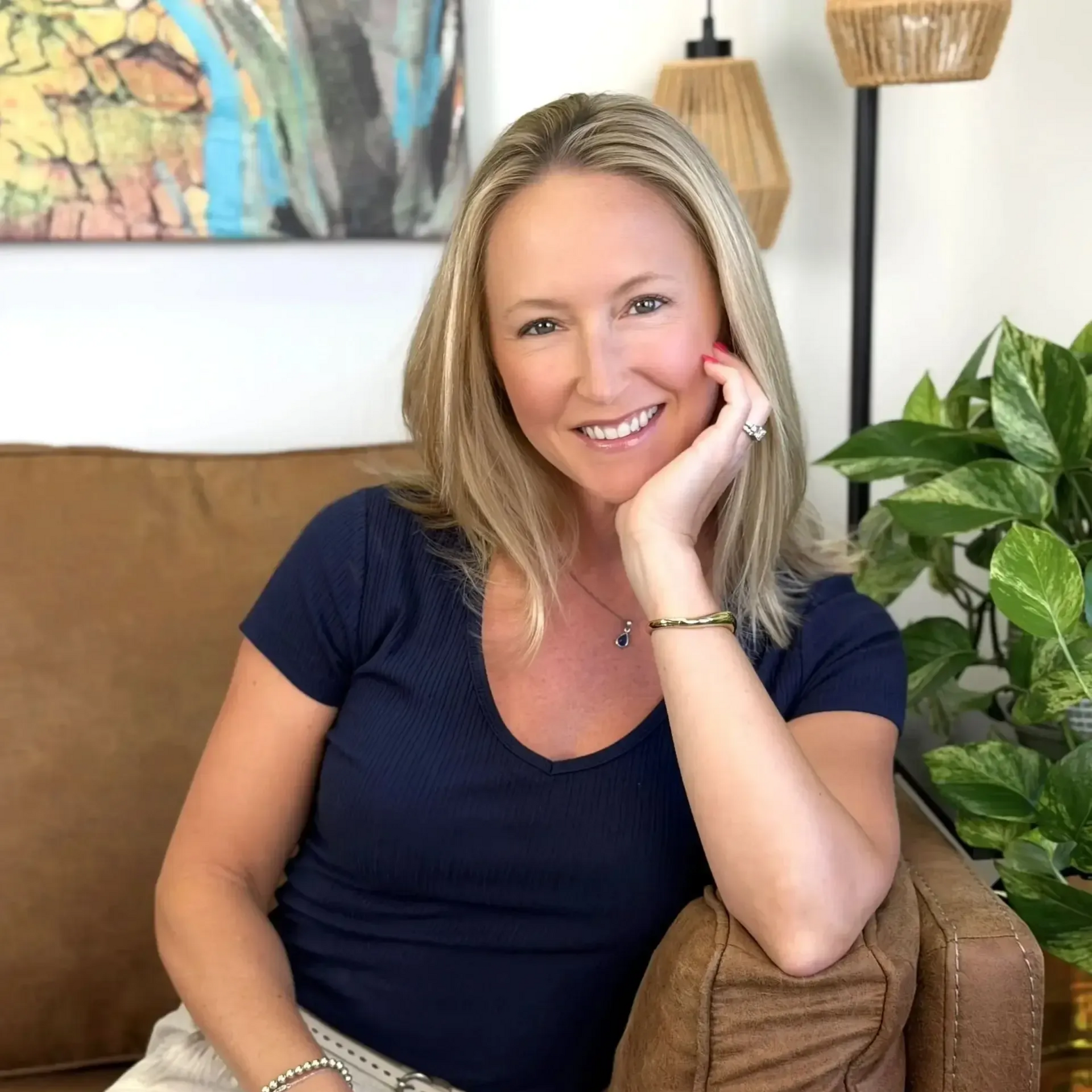 Smiling woman in a navy shirt reclining on a couch beside a green plant