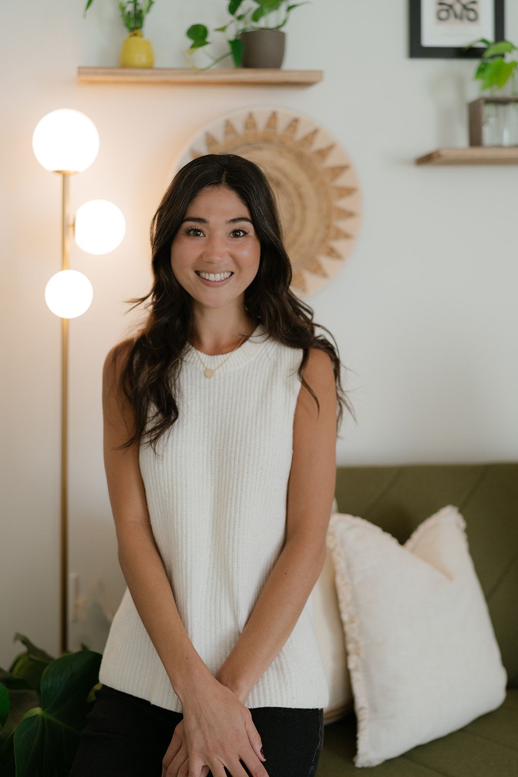 Woman standing in a living room beside a floor lamp, smiling, with a couch and wall decor behind her