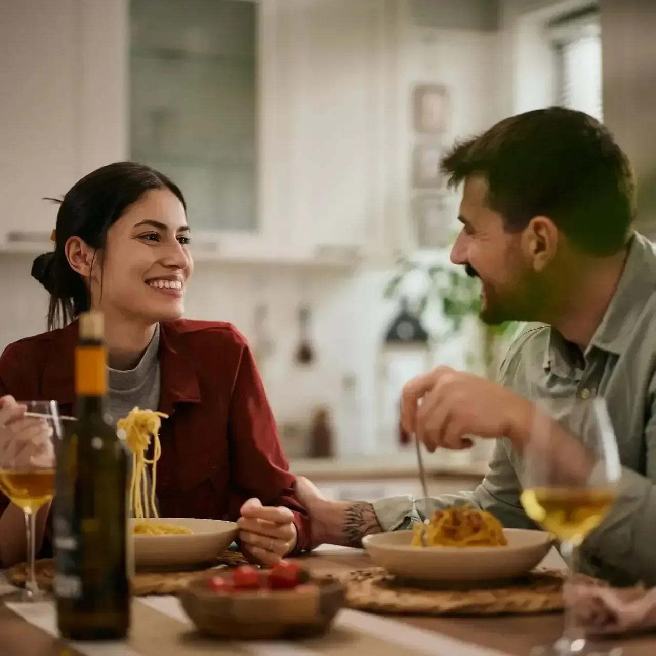 Two people smiling at a dinner table, talking over a meal with wine glasses and a bottle on the table