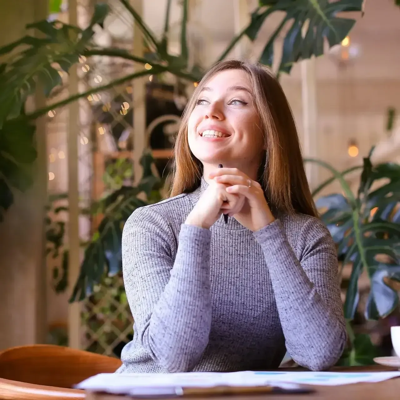 Smiling woman in a gray sweater seated at a table in a plant-filled room, looking upward thoughtfully