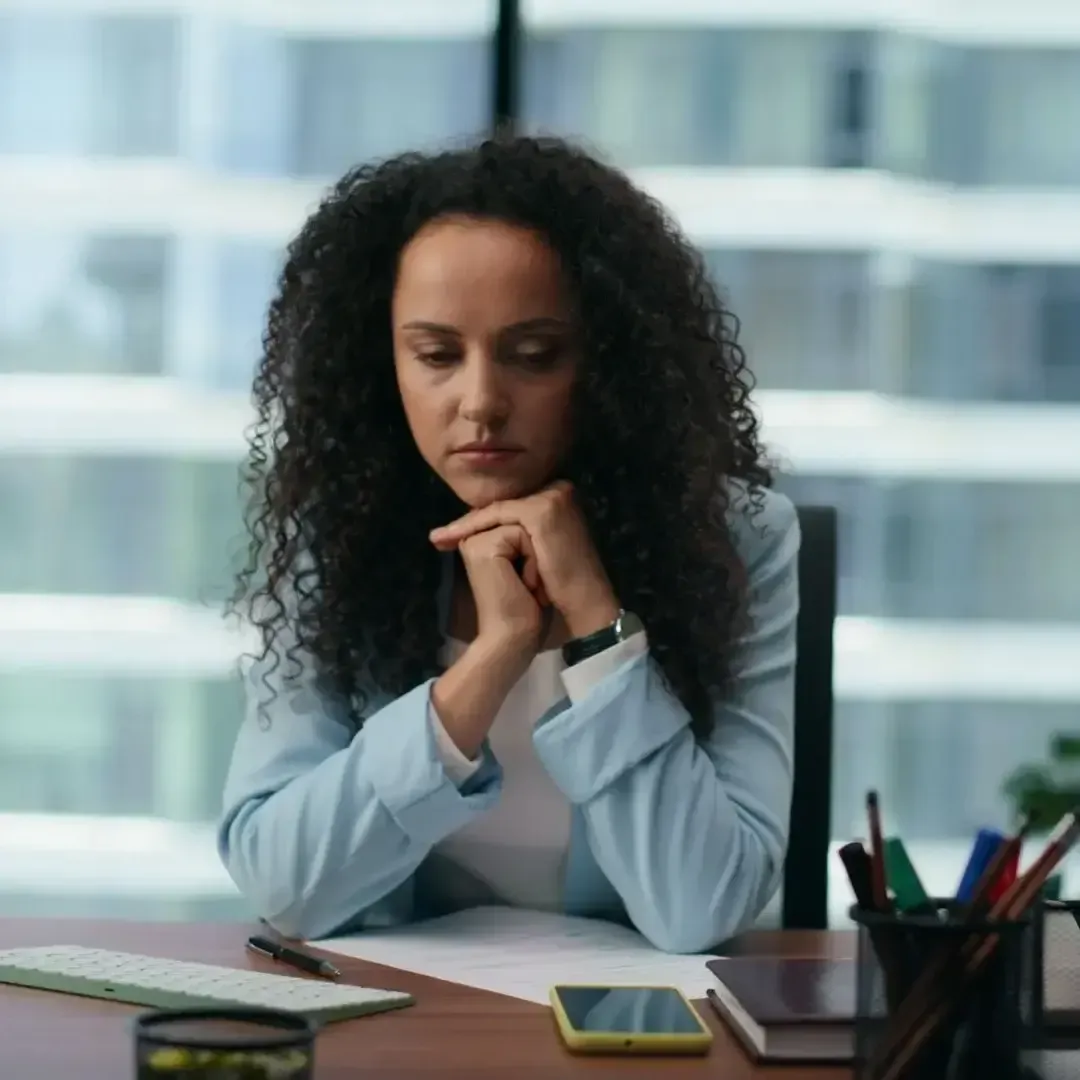 Woman in a light blue shirt sitting at a desk, looking down thoughtfully with hands clasped.