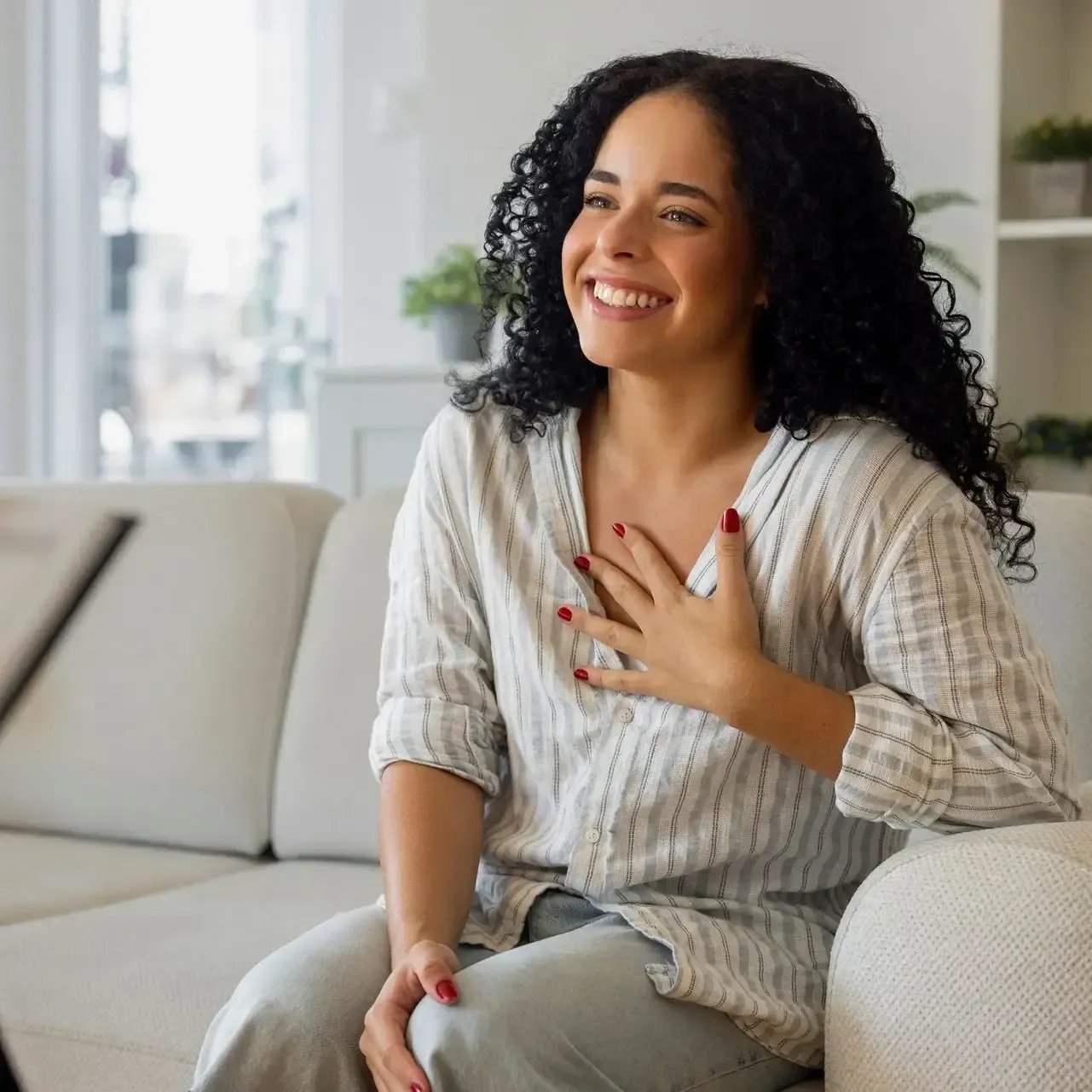 Woman smiling on a sofa, hand on chest, in a bright living room