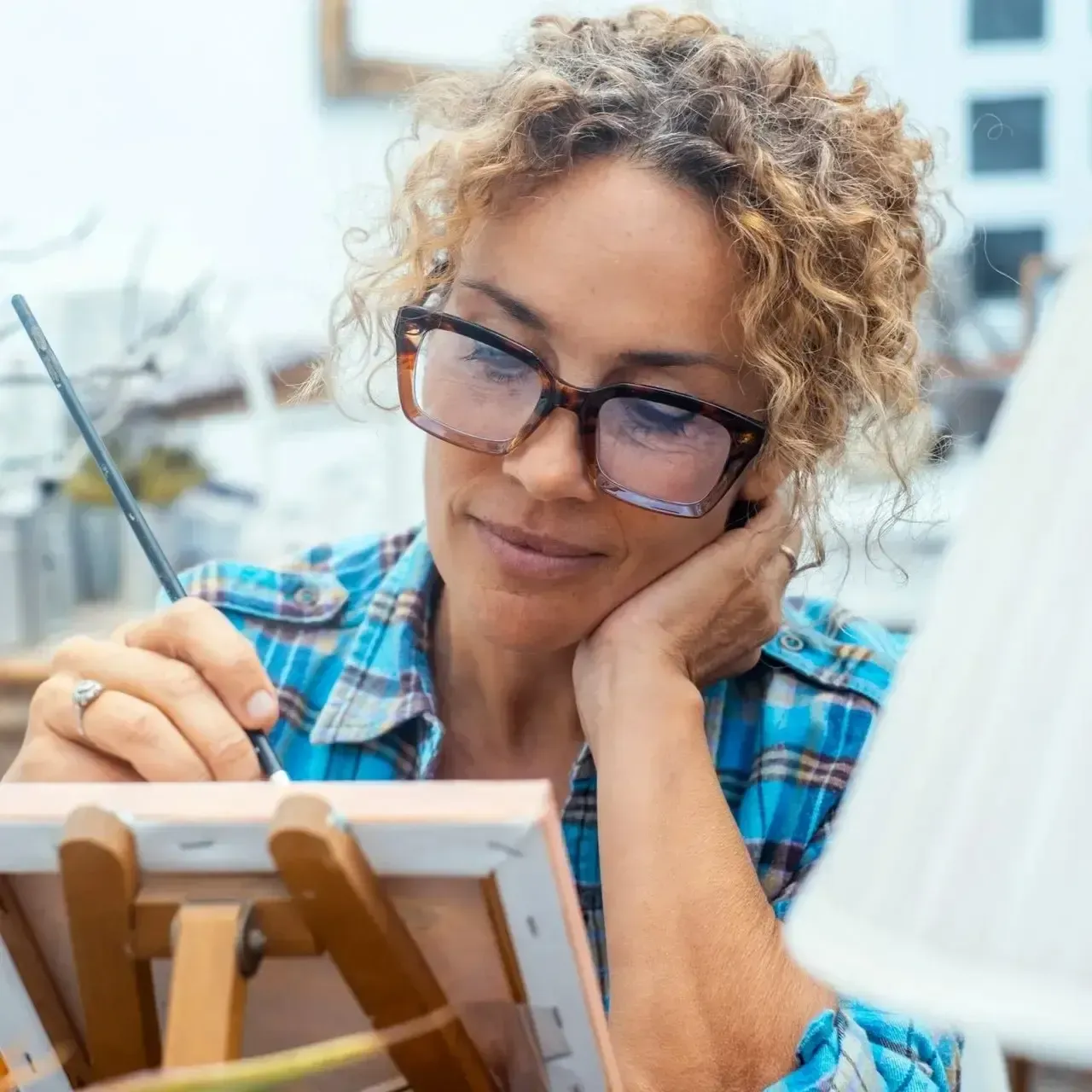 Person sketching on a canvas in a bright studio, wearing glasses and a blue plaid shirt.