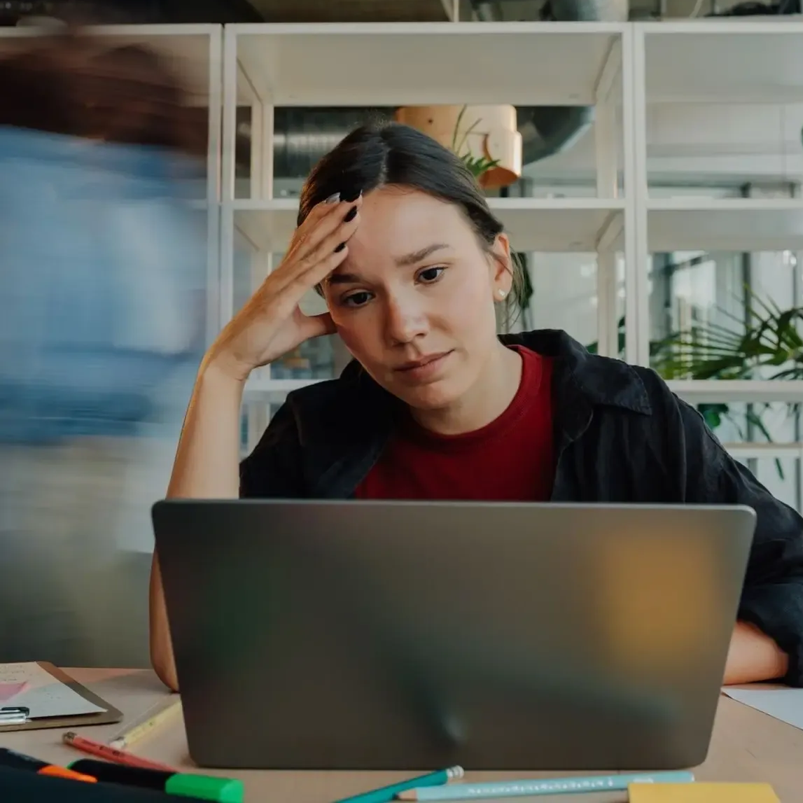 Person sitting at a laptop, holding their head at a desk in a bright office, looking stressed.