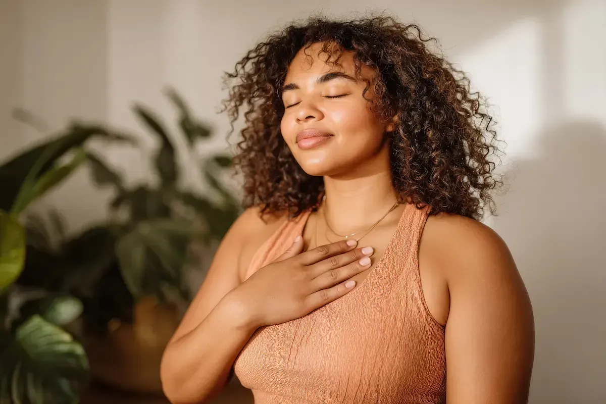 Woman with curly hair, eyes closed, hand on chest, wearing a peach sleeveless top in a soft-lit room
