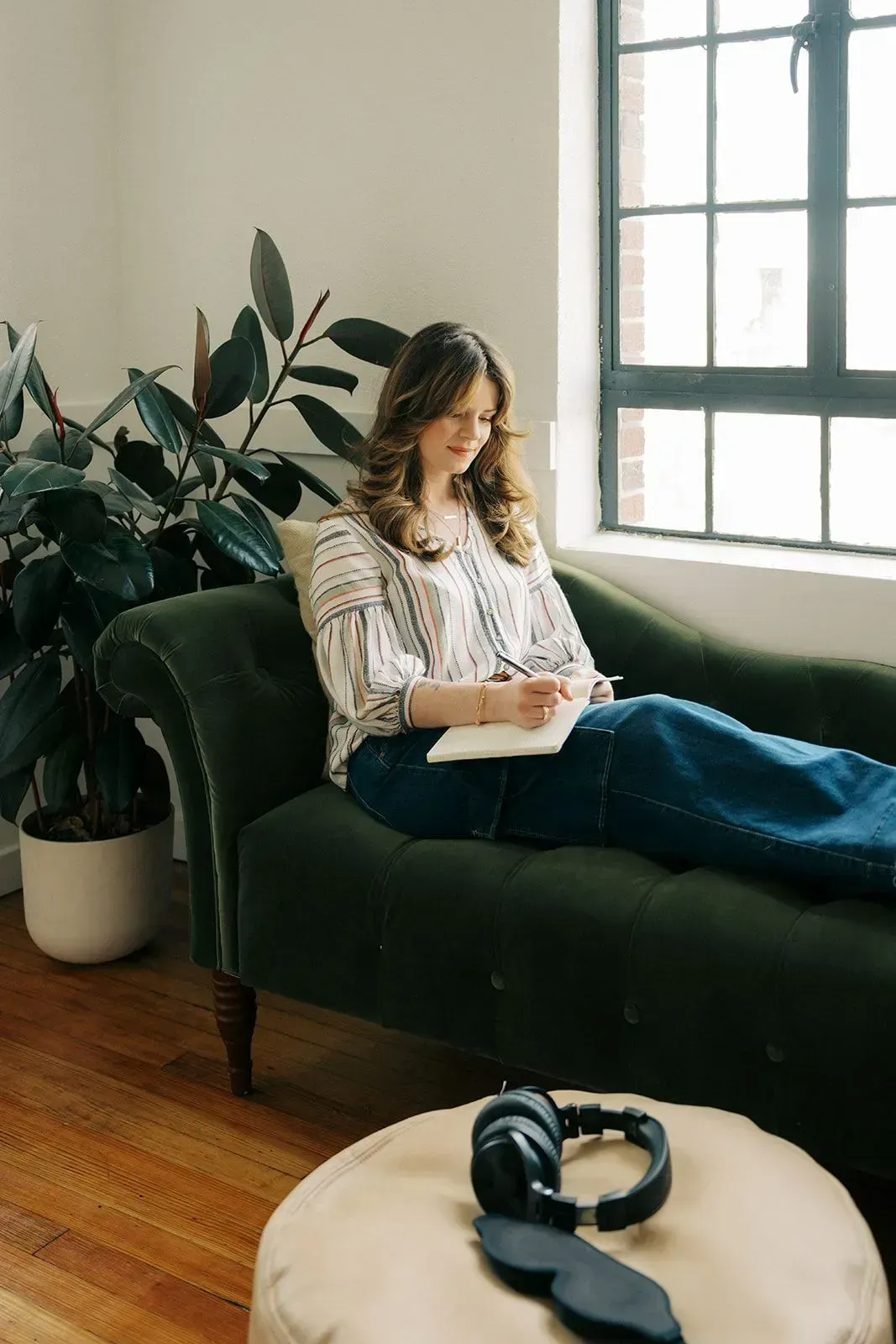 Jennifer Lancaster on a green chaise by a window, reading a book with headphones on a nearby table.
