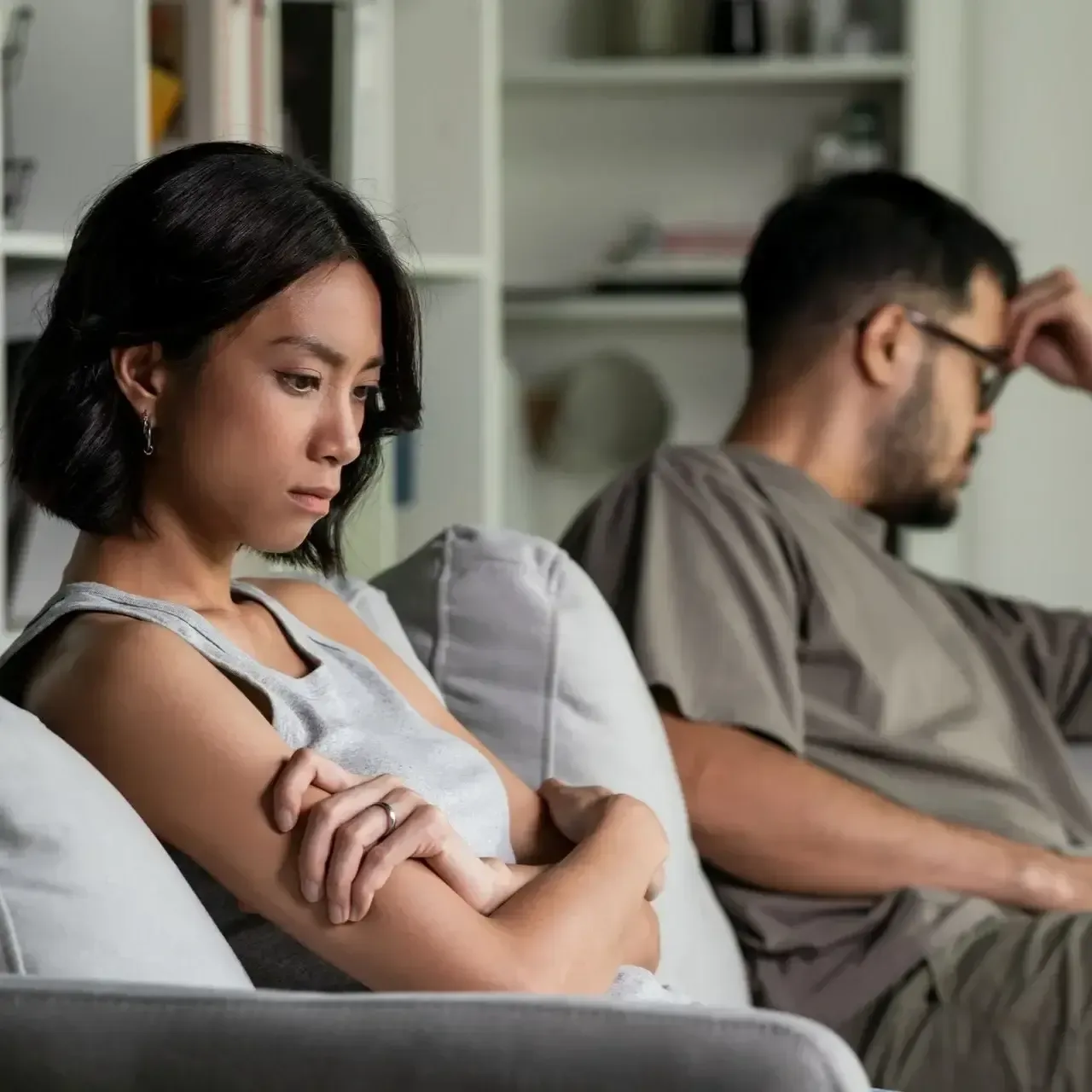 Two people sit apart on a couch, looking away in a tense living room scene.