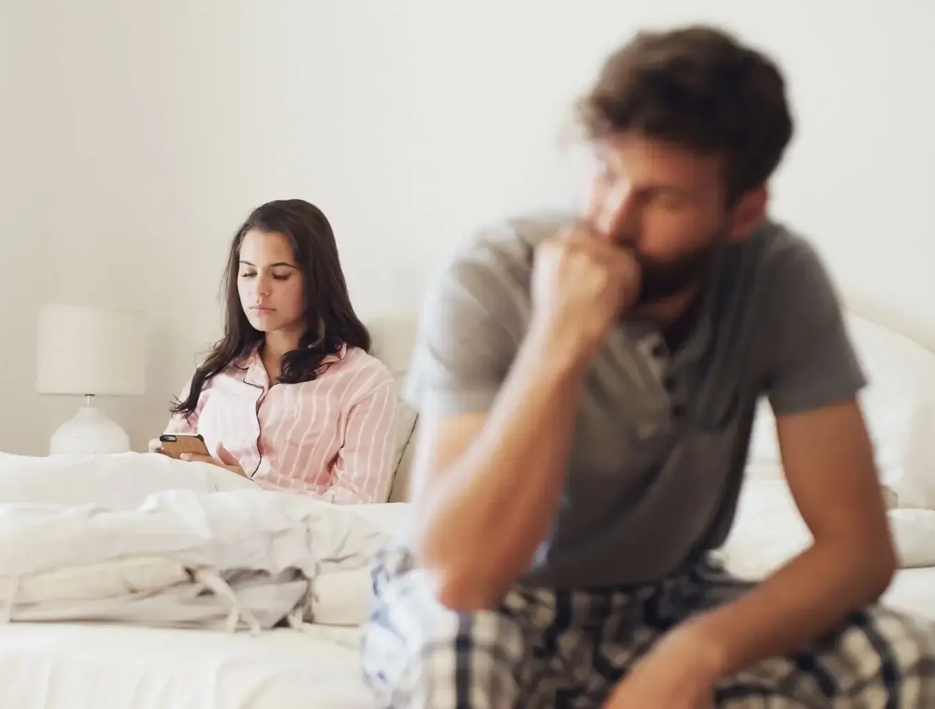 Couple sitting apart on a bed, one looking at a phone, the other with head in hand, in a tense room
