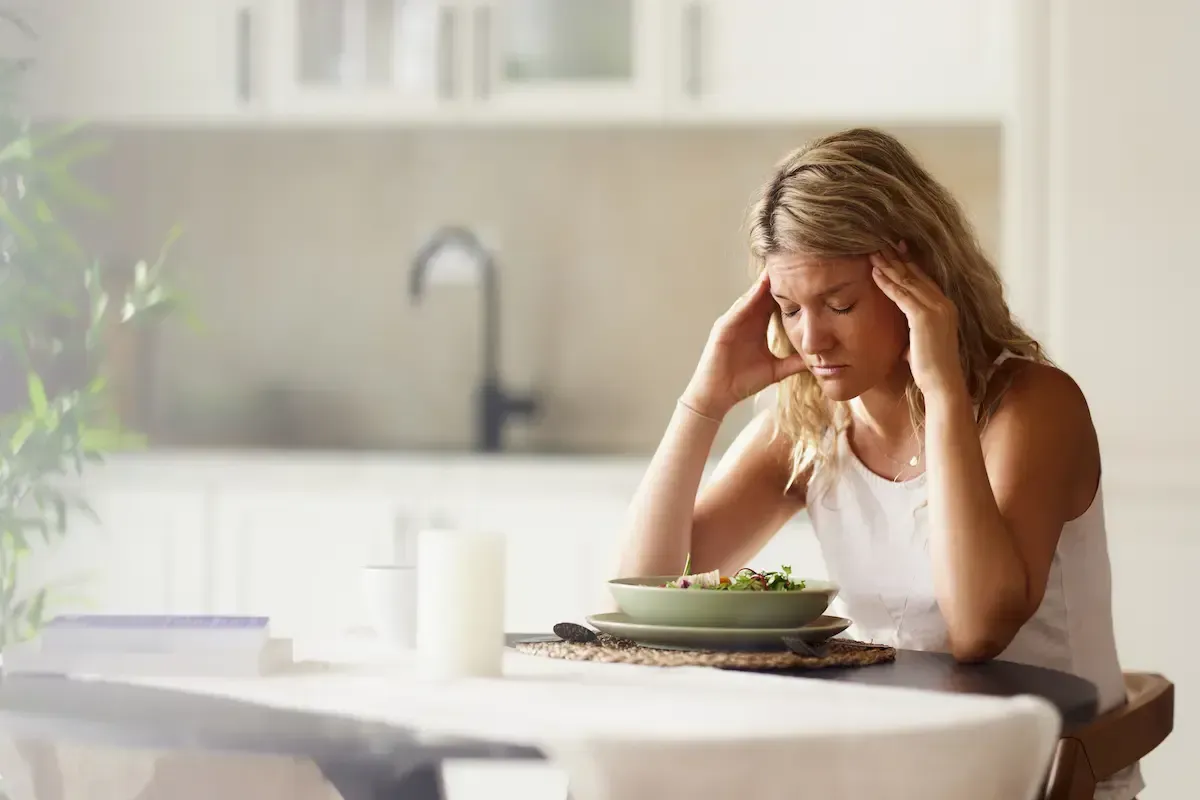 Person sitting at a table with head in hands beside a plate of food, looking stressed in a bright kitchen