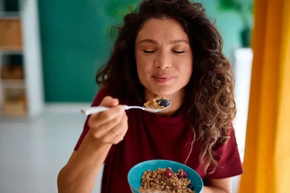 Woman eating cereal from a blue bowl with a spoon, eyes closed in a cozy kitchen.