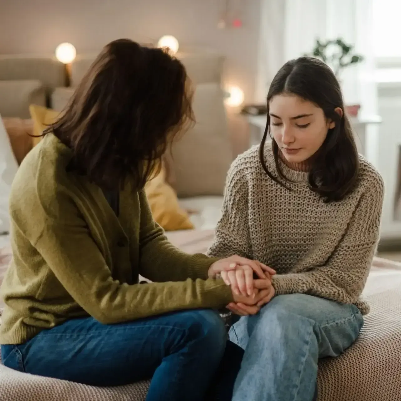Two women sit on a sofa, holding hands in a warm, softly lit room.