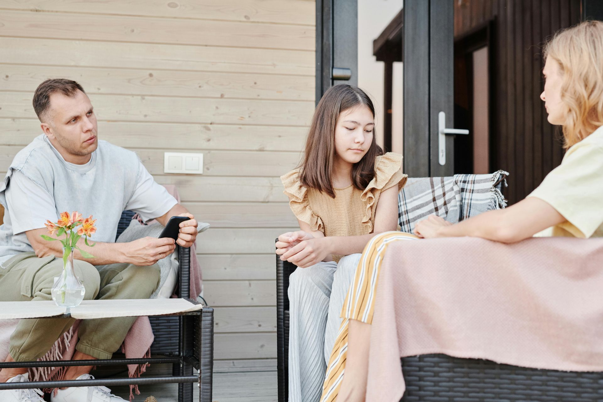 Three people sitting and talking on a patio with wicker chairs and a small table of flowers