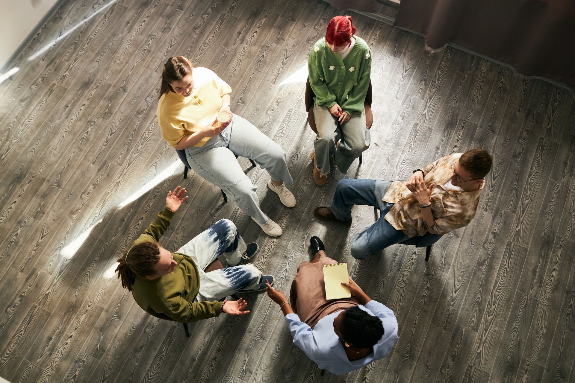 Five people sit in a circle on a wooden floor, seen from above, in casual conversation.