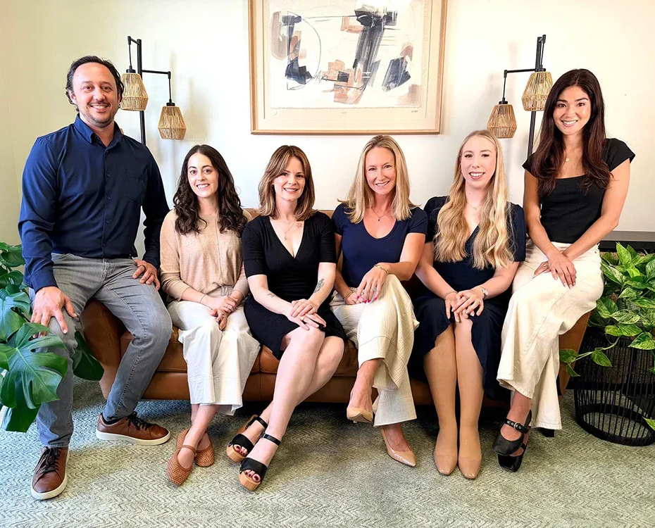 Six people seated and standing in a bright living room, smiling at the camera.