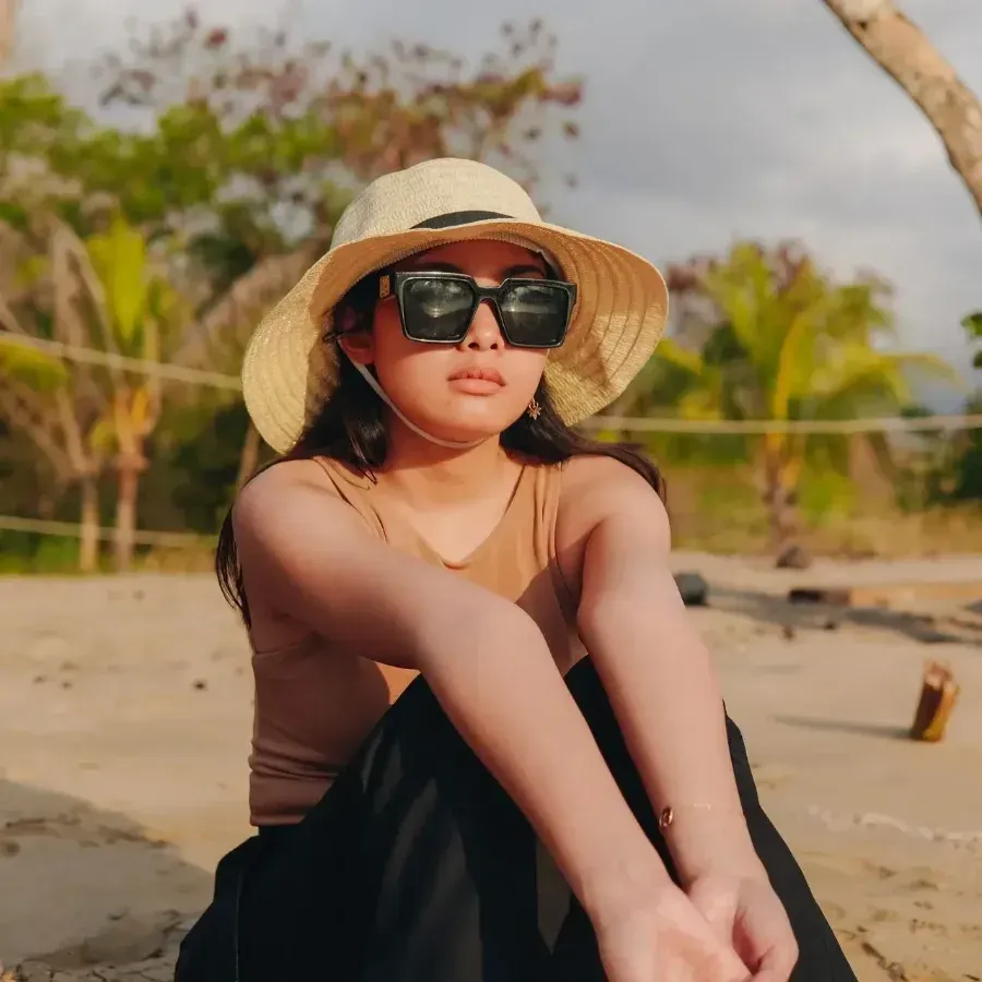 Woman in sunglasses and sunhat sitting on a sandy beach with palm trees in the background