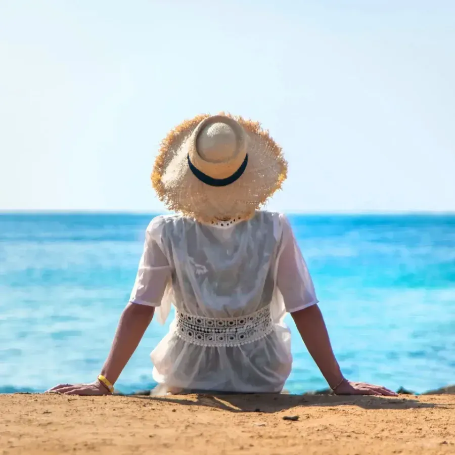 Person in a sunhat sits facing the ocean on a sandy beach under a clear blue sky
