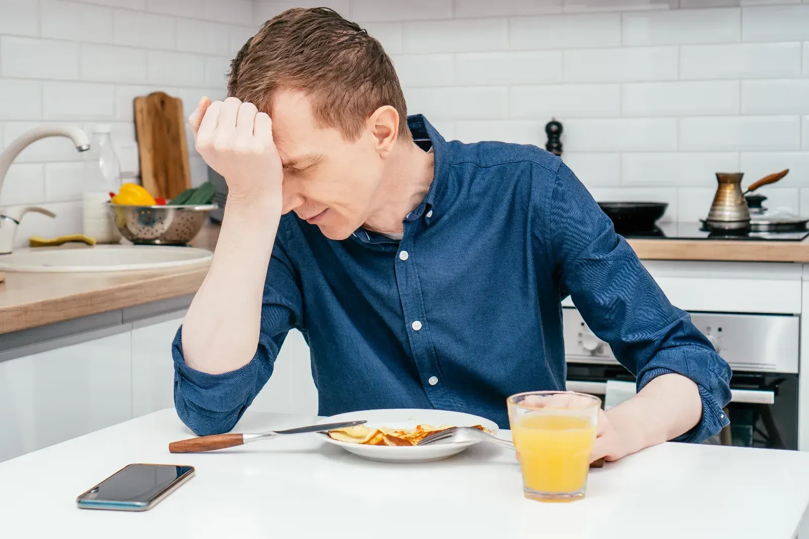 Person sitting at a table with head in hands beside a plate of food, looking stressed in a bright kitchen