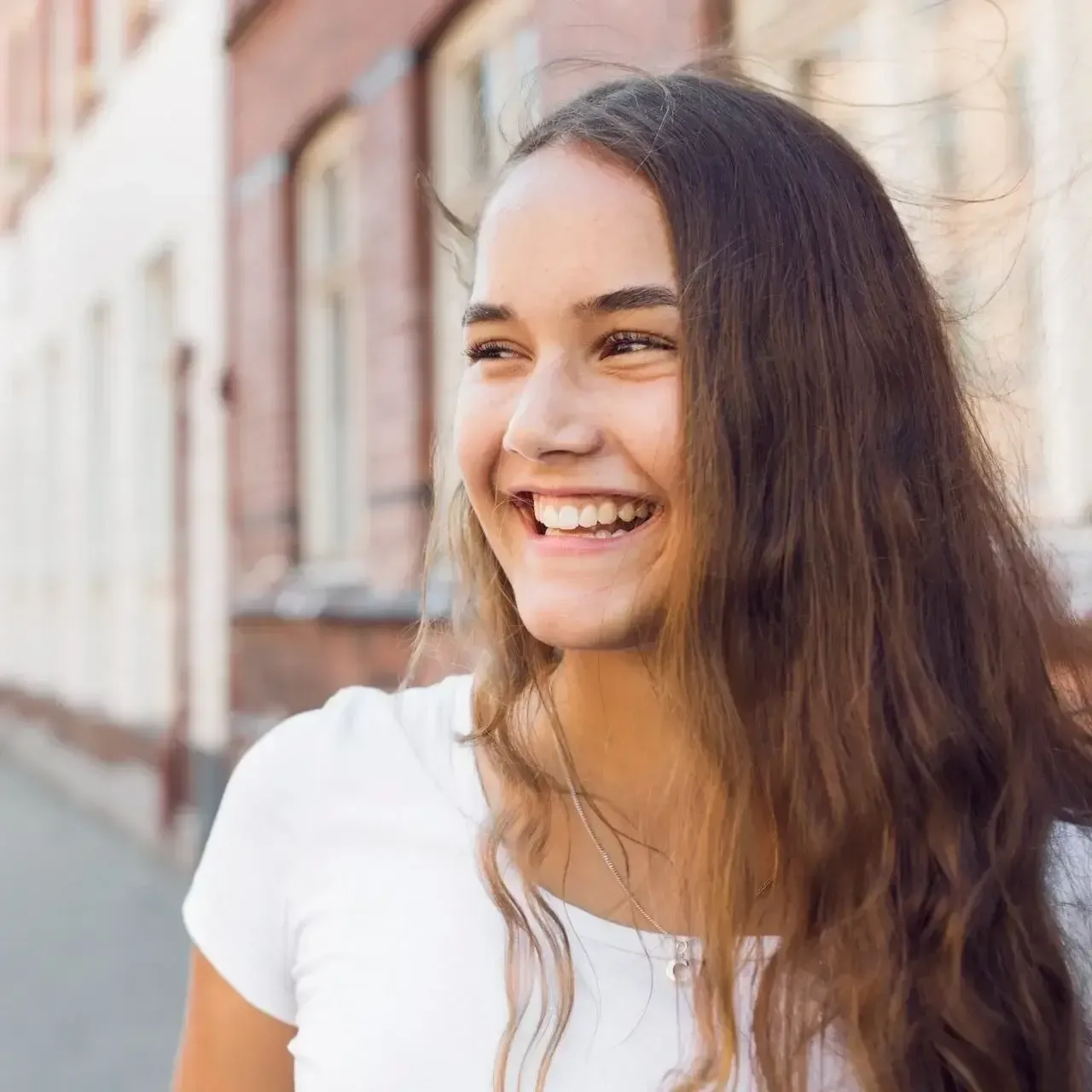 Smiling woman in a white shirt standing on a sunlit street beside brick buildings
