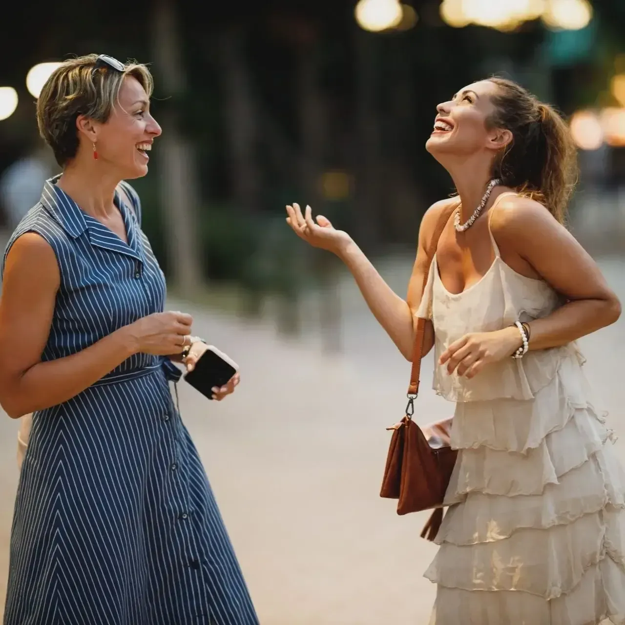 Two people laughing and chatting outdoors, one in a blue dress and one in a cream ruffled dress.