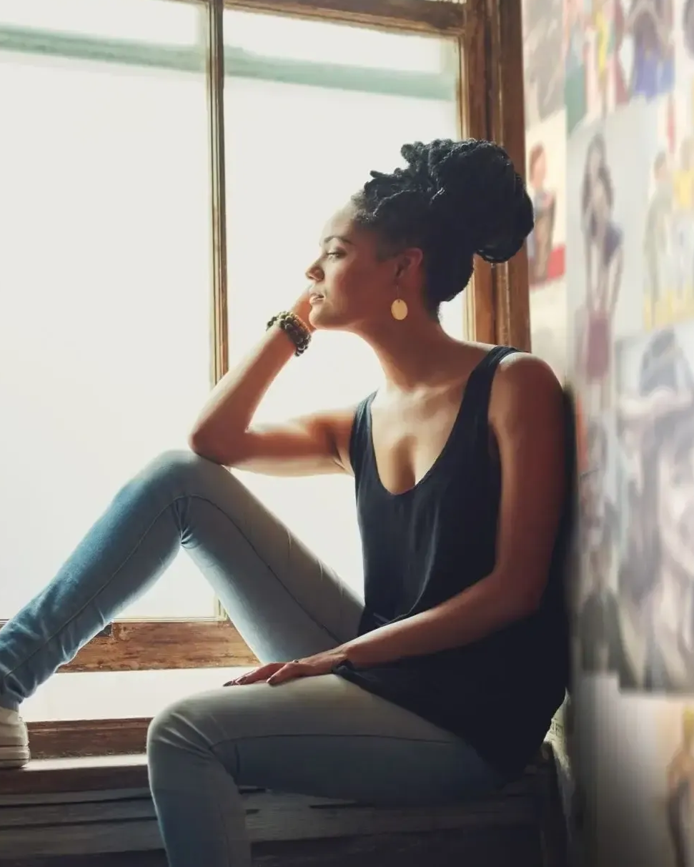 Person sitting by a window, gazing outside in a black tank top and jeans.
