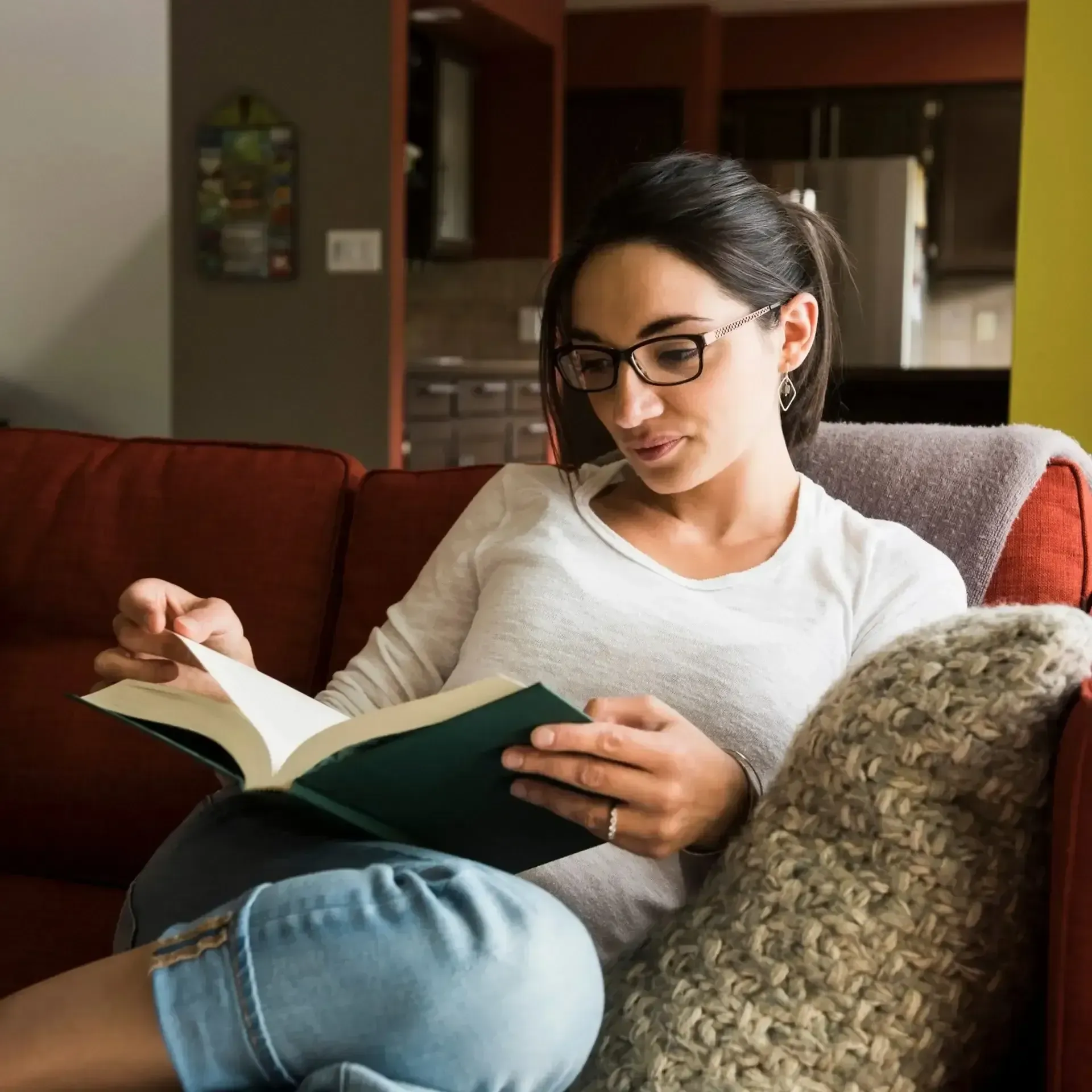 Woman reading a book on a couch in a living room, wearing glasses and a white shirt.