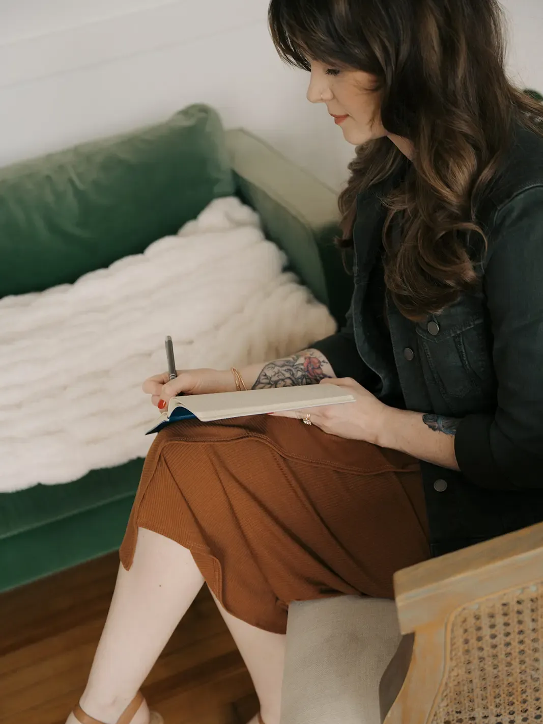 Woman sitting on an olive sofa in a bright room, facing left, with plants and a wall lamp nearby